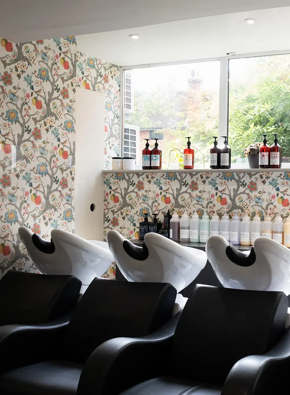 Interior of Albert Fieldshair salon in Oxfordshire with three black reclining shampoo chairs, white sinks, floral wallpaper, a window with natural light, and shelves with hair products and conditioners.
