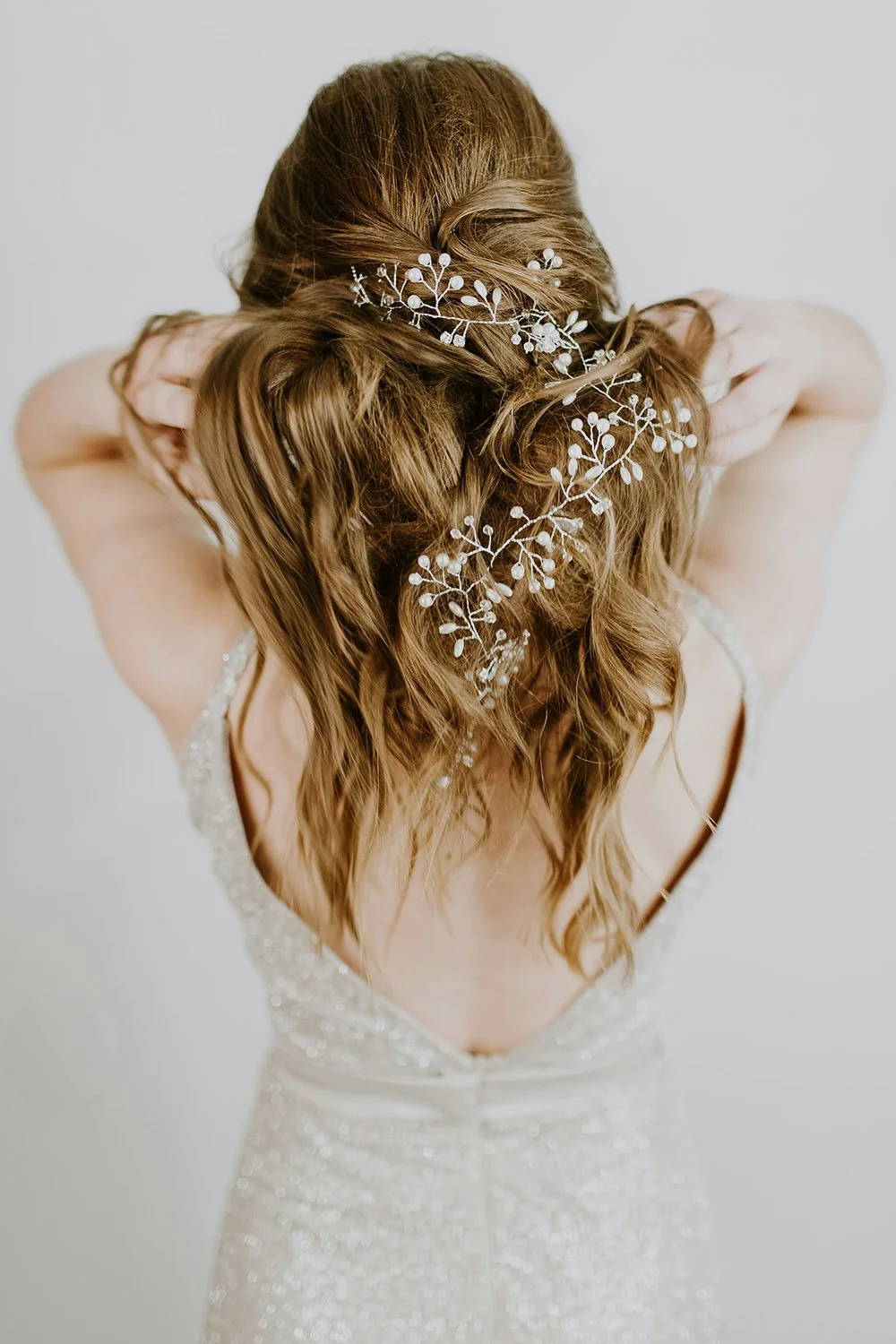 A woman with wavy, reddish-brown hair styled in loose waves, decorated with a delicate silver hairpiece with floral and leaf design, wearing a sparkly, backless wedding dress.