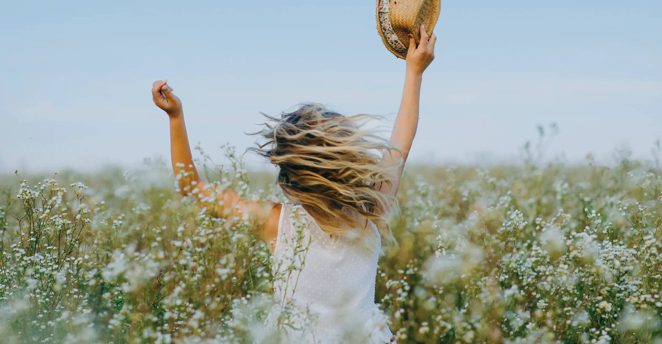 Back view of a woman with blonde wavy hair running joyfully through a wildflower field, holding a straw hat, her hair styled with natural movement at Albert Fields salon