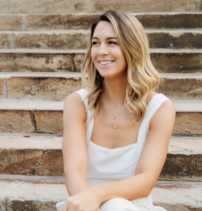 A smiling woman with blonde hair sitting on stone steps, wearing a white sleeveless top and a delicate necklace.