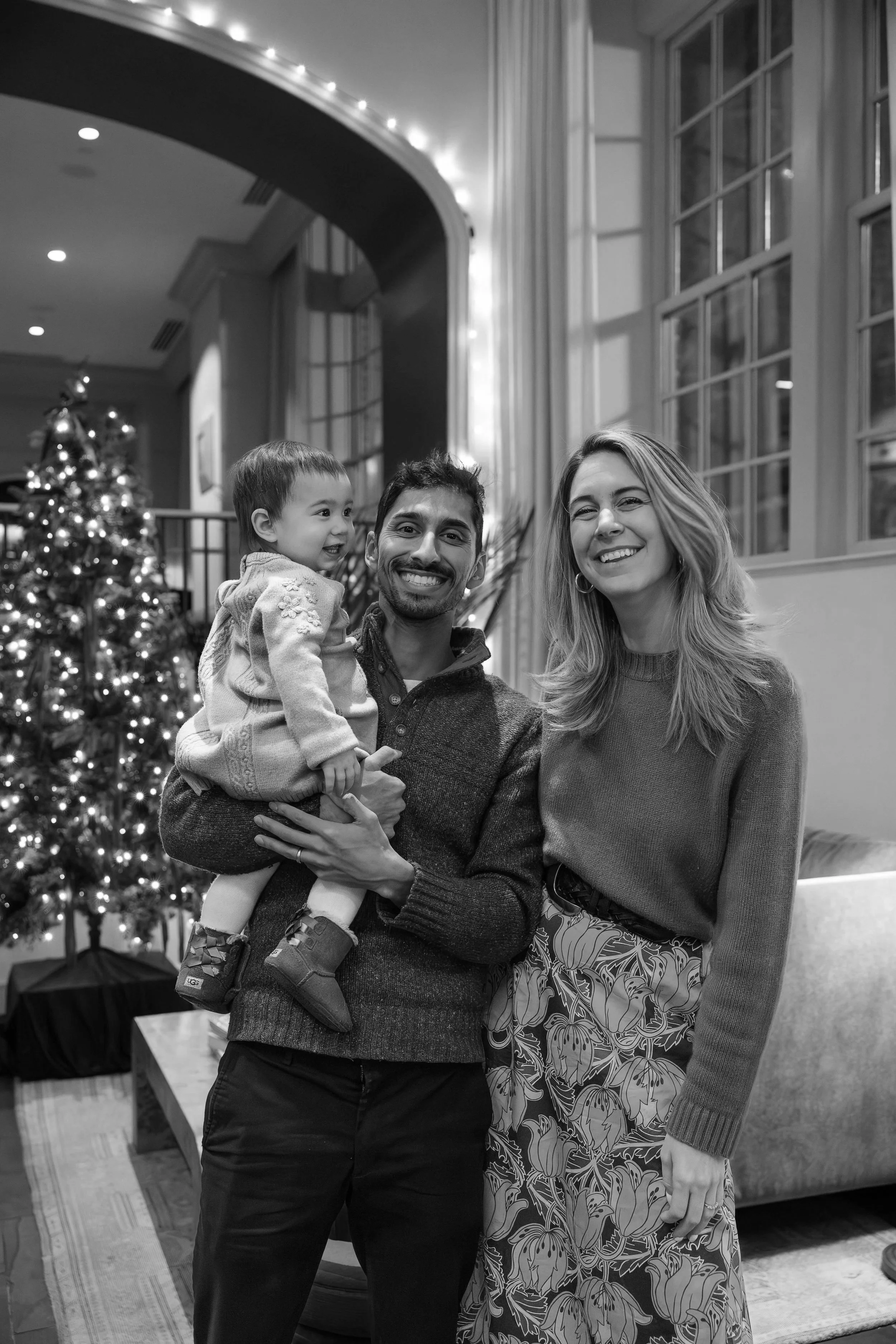 A happy family of three, including a man, woman, and a young girl, is posing together indoors during Christmas time. There is a decorated Christmas tree in the background.