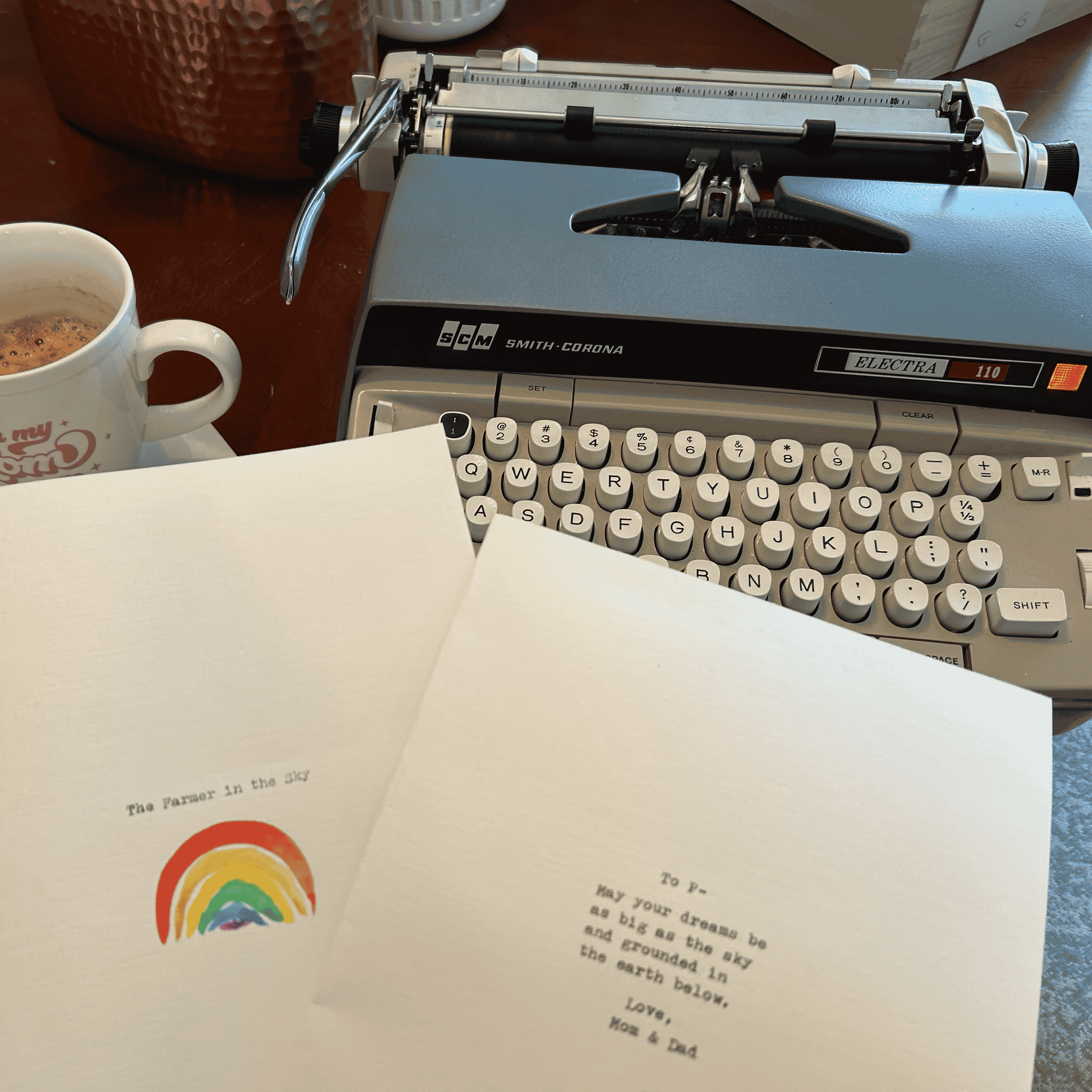 A vintage typewriter on a wooden table with sheets of paper, a cup of coffee, and a rainbow illustration, with part of a colored chart in the background.