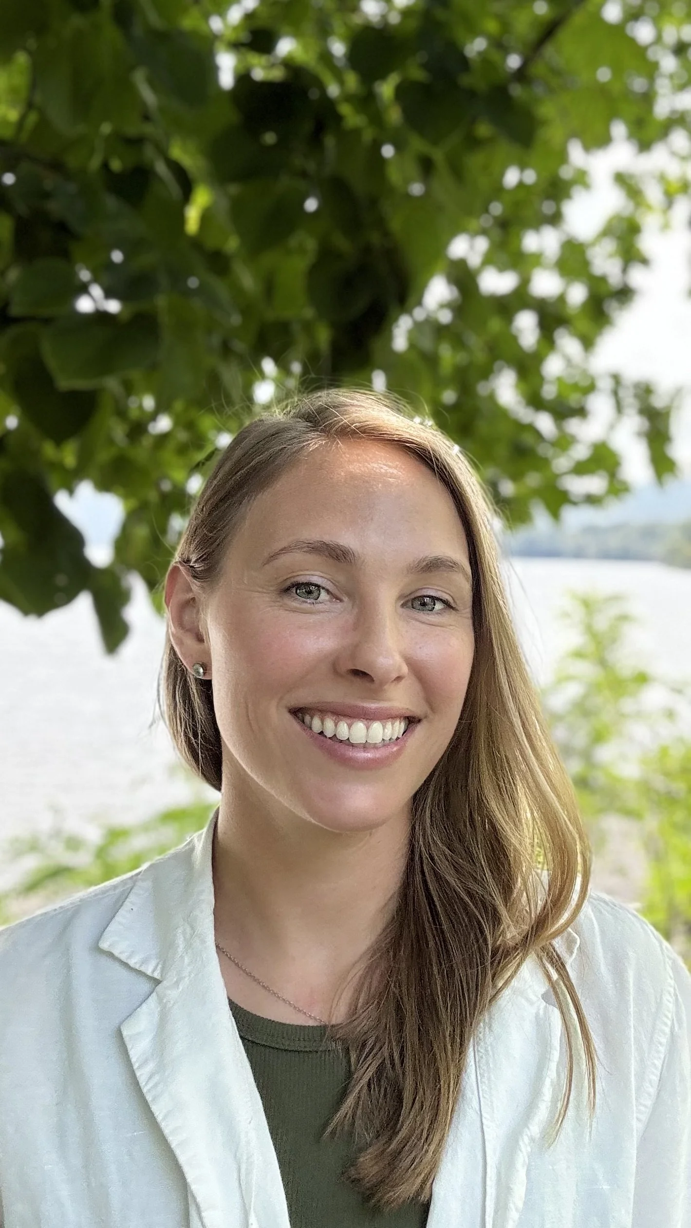 A woman with long, light brown hair and a wide smile standing outdoors near a lake with green trees and foliage in the background.