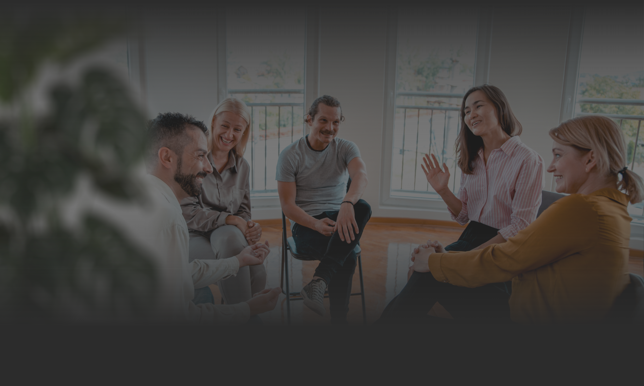 Group of six people sitting and talking in a bright room with large windows, some smiling and engaging in conversation.