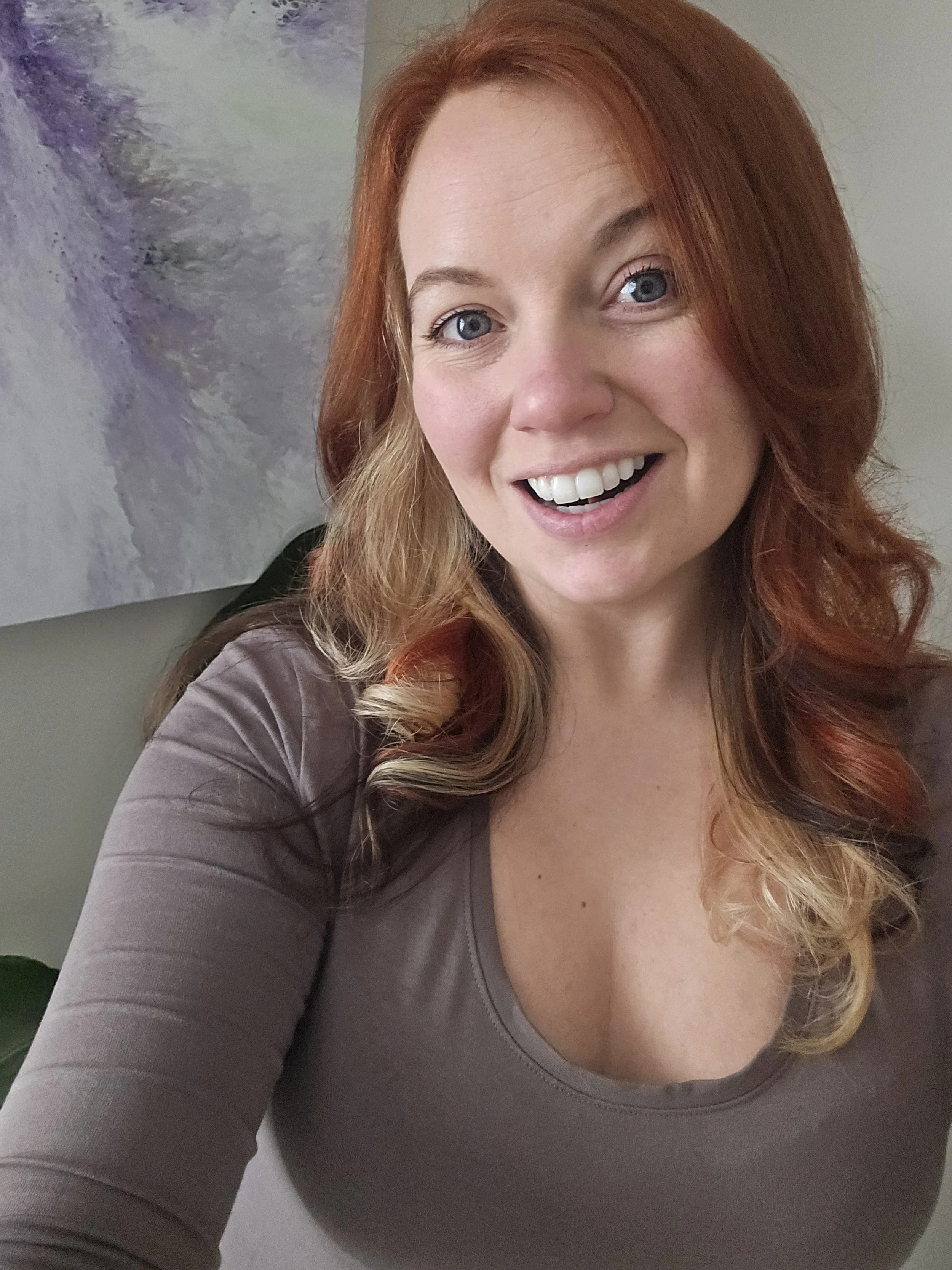 A woman with wavy red hair and blue eyes smiling at the camera, wearing a gray top. Part of an abstract painting is visible in the background.