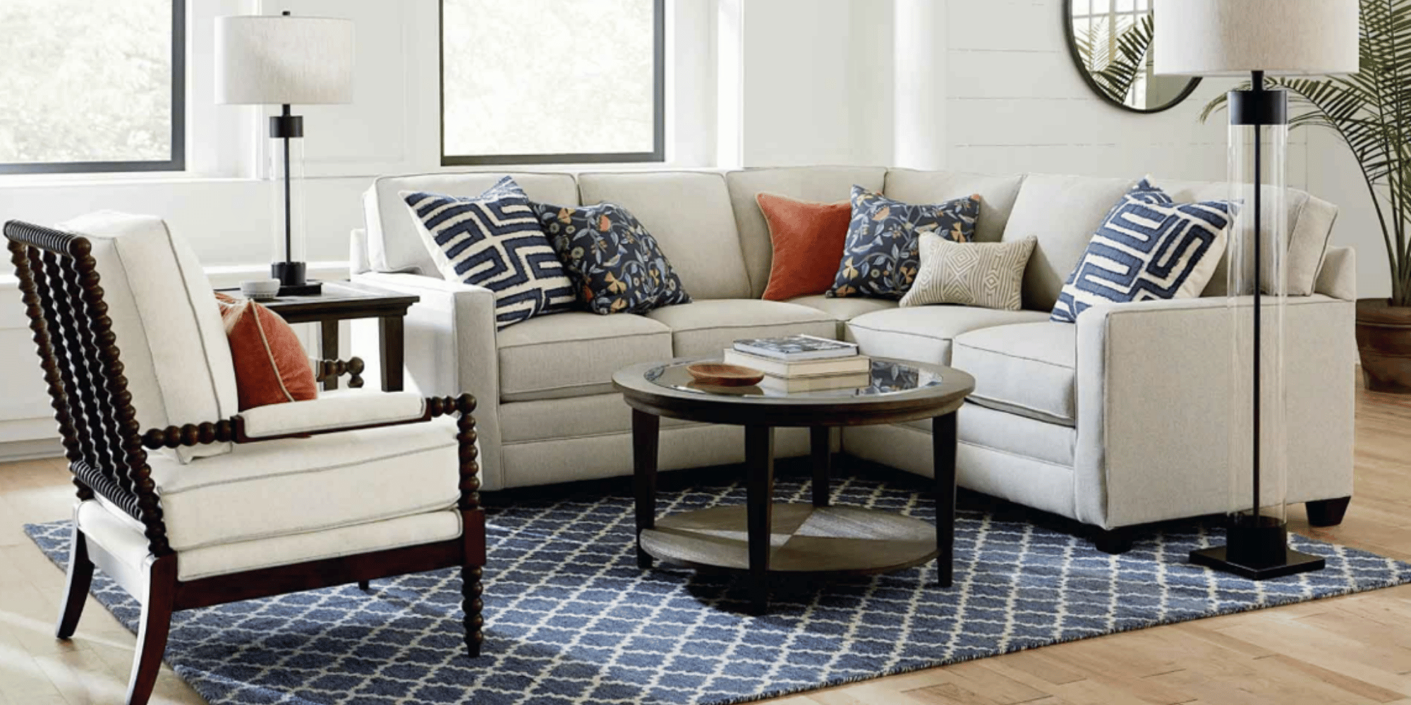 Living room with white sofa and armchair, decorated with patterned cushions in blue, beige, and rust colors. A wooden coffee table and side table, black floor lamps, and a blue patterned rug complete the setting. Windows in the background and a mirro