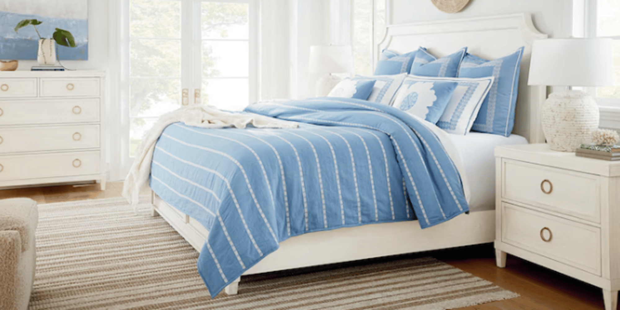 A bedroom with a white bed frame, blue striped bedding, and several matching blue pillows. There are white nightstands on either side of the bed with lamps, and a dresser with a plant and artwork on top. Large windows let in natural light.