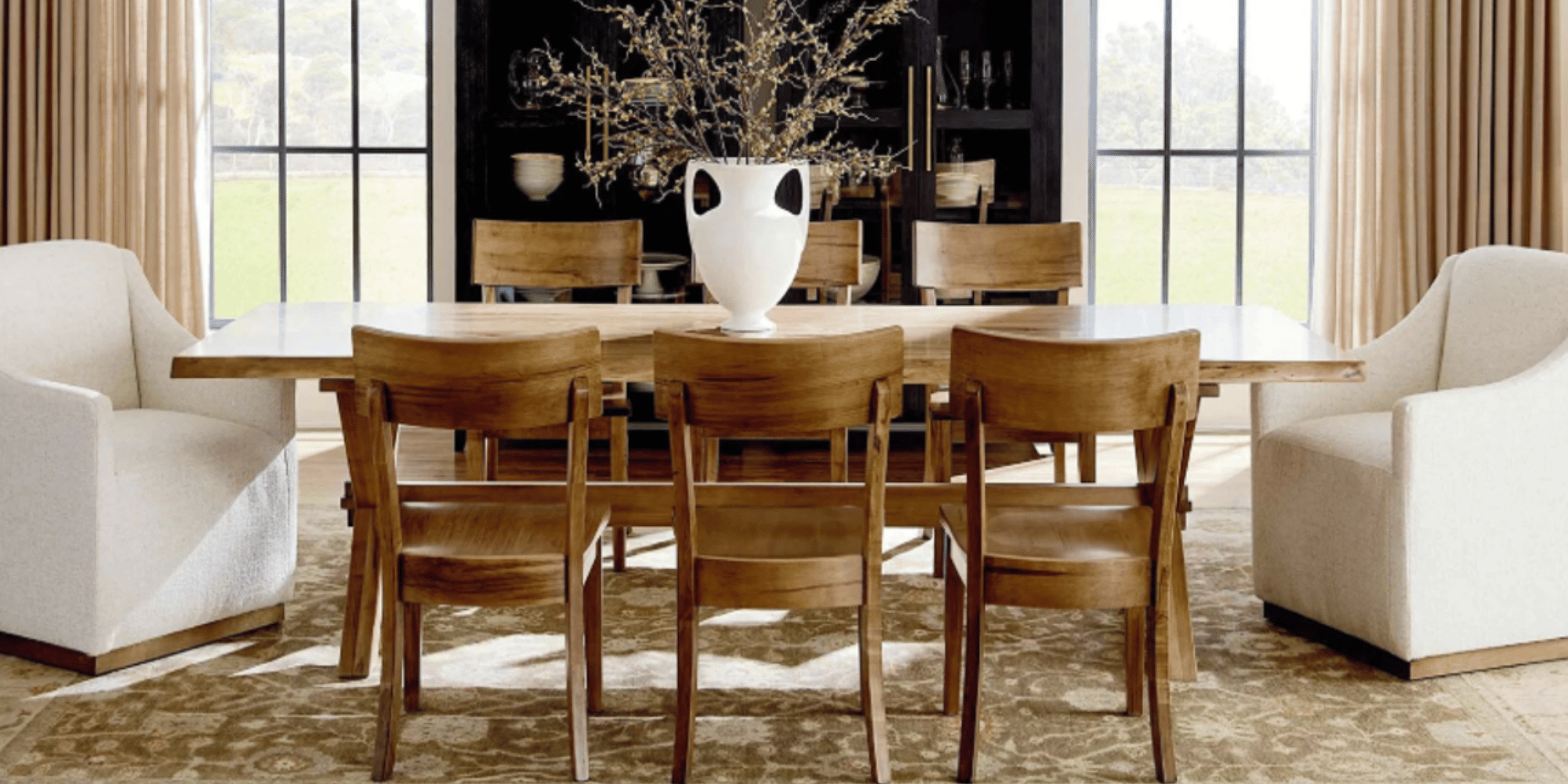 A modern dining room with a large wooden dining table surrounded by a combination of eight wooden chairs and two white upholstered armchairs. There is a decorative white vase with dried branches on the table. In the background, there is a black wall 
