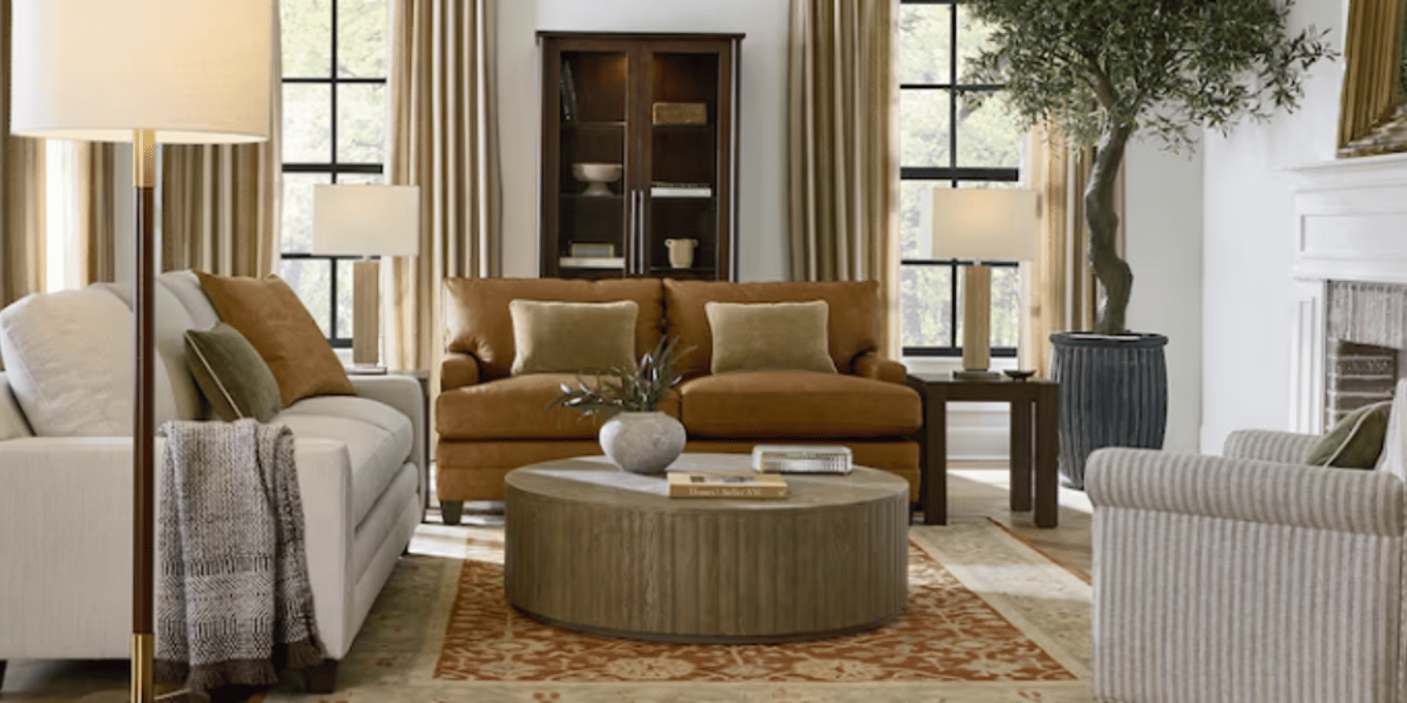 A cozy living room with a mix of white and tan sofas, a round wooden coffee table, tall lamps, a dark wood cabinet, large windows with beige curtains, a potted olive tree, and a decorative fireplace.