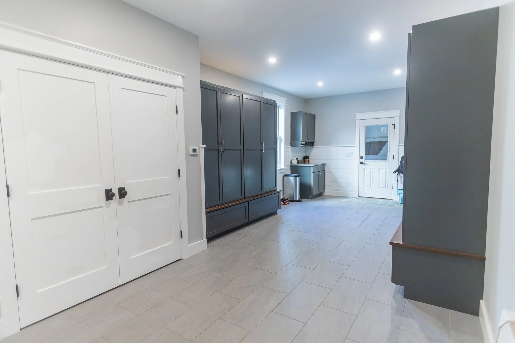 Interior view of a modern kitchen with white walls, gray cabinetry, a small countertop area near a window, and a door leading outside. Recessed ceiling lights illuminate the space, and the floor is tiled.