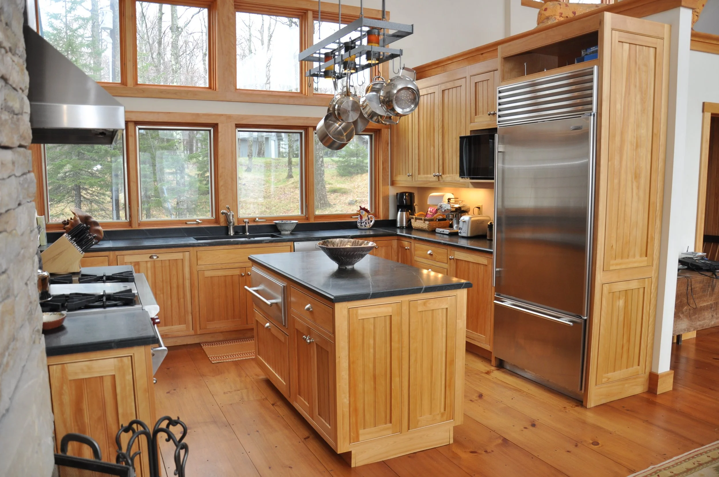 A well-lit kitchen with large windows showing trees outside. Wooden cabinets, a kitchen island, and modern appliances including a stainless steel refrigerator. Pots and pans hang from a ceiling rack above the island.