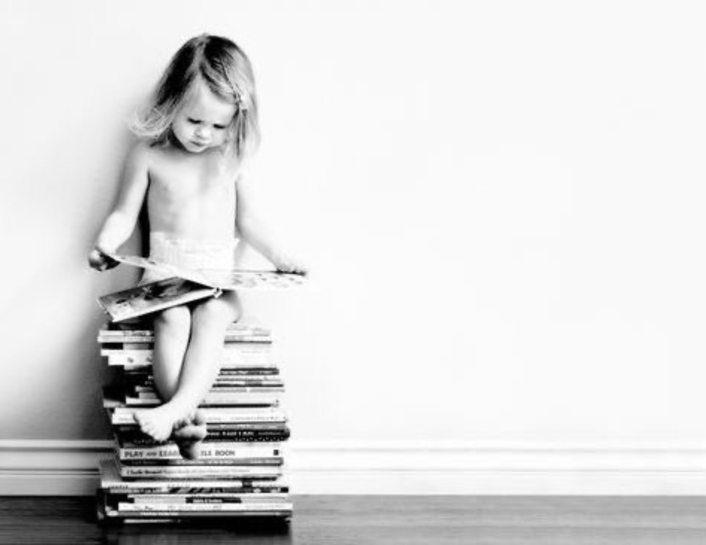 Niño pequeño sentado sobre una pila de libros, leyendo un libro en blanco y negro.