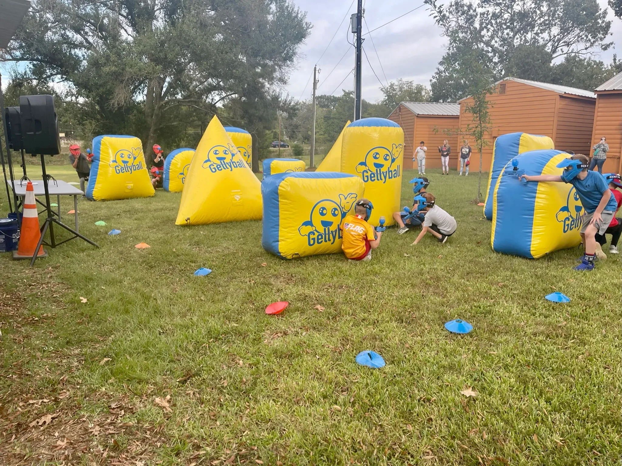 People playing a game of paintball outdoors with yellow and blue inflatable obstacles labeled 'Gellyball,' some wearing helmets and protective gear, on a grassy field with trees and a building in the background.