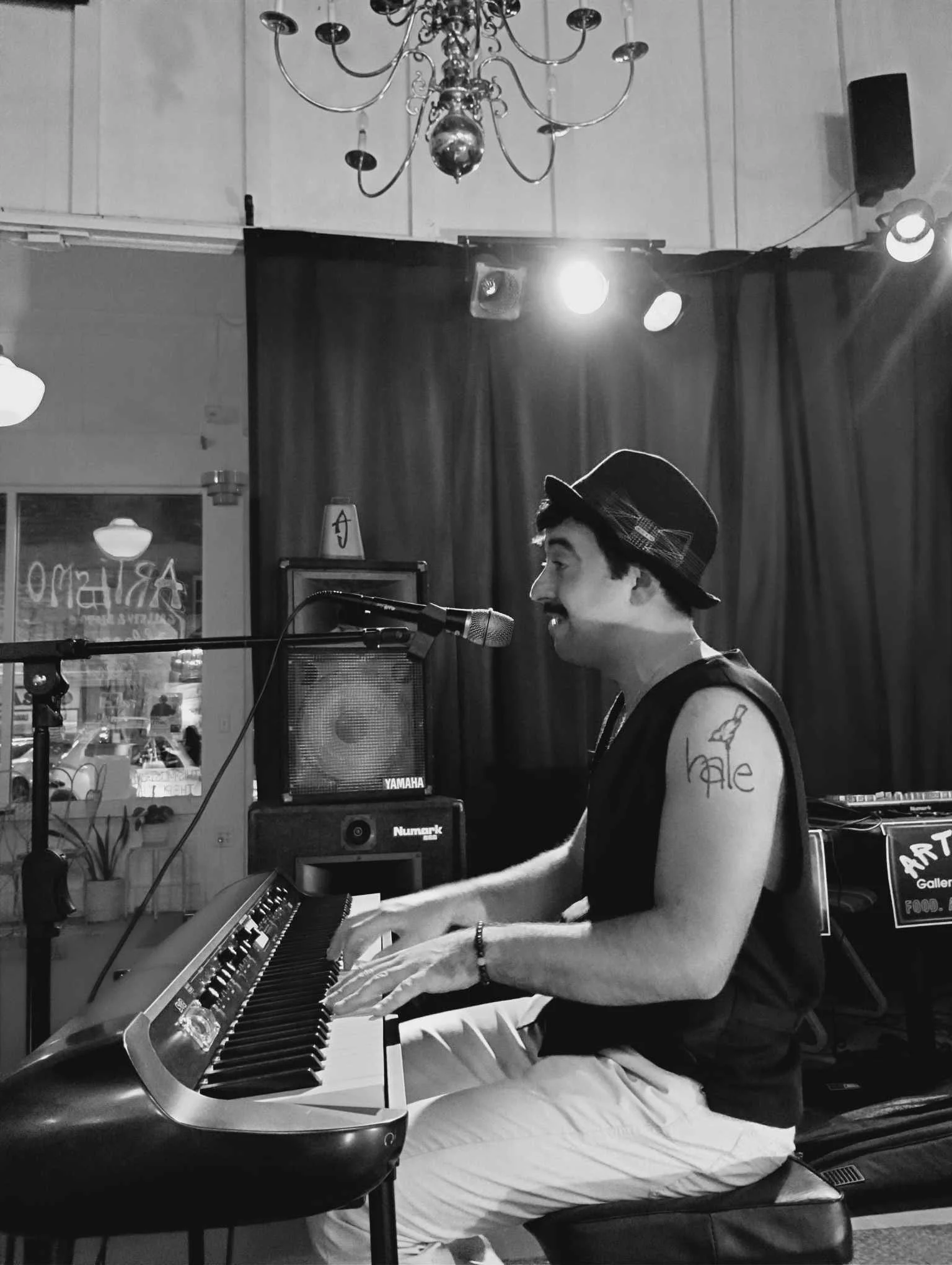 A person playing a keyboard piano in an indoor setting with stage lighting and a curtain backdrop. The person is wearing a black hat with a checked band, a sleeveless shirt, and white pants. They have a tattoo on their left arm that says 'bale' with a bird. There are musical equipment and speakers visible in the background.