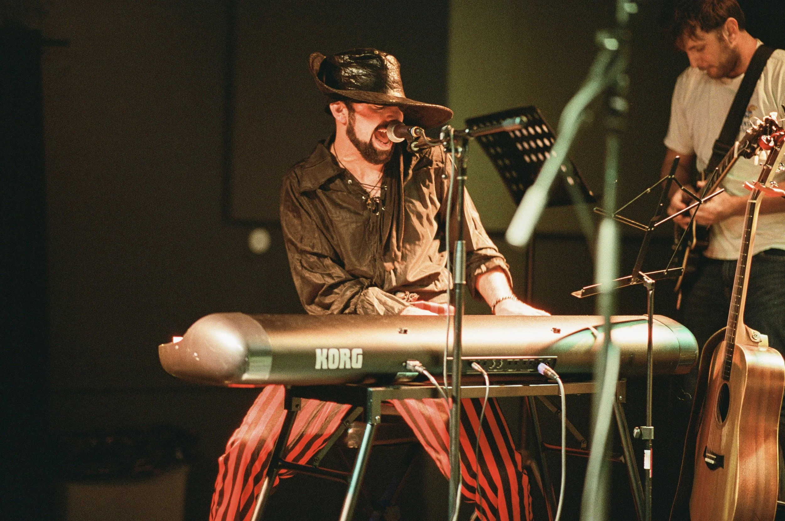 Joey Crofoot singing and playing keyboard on stage, wearing a pirate hat and striped red and black pants, with another musician playing guitar nearby.