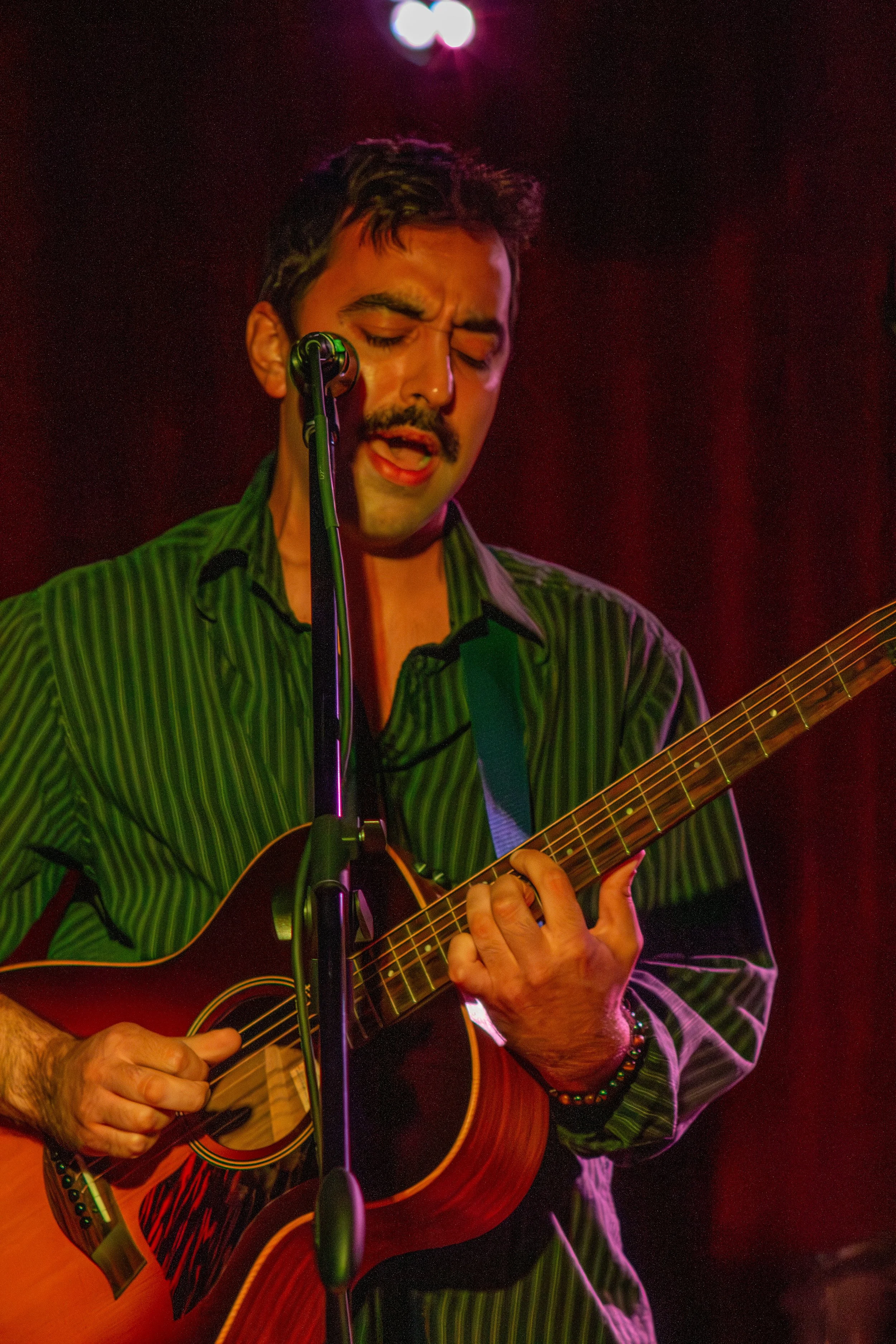 Joey is singing passionately while playing an acoustic guitar on stage. He is wearing a green striped shirt and has a colorful beaded bracelet on his left wrist. The stage is dimly lit with a red background, and there is a microphone in front of him.