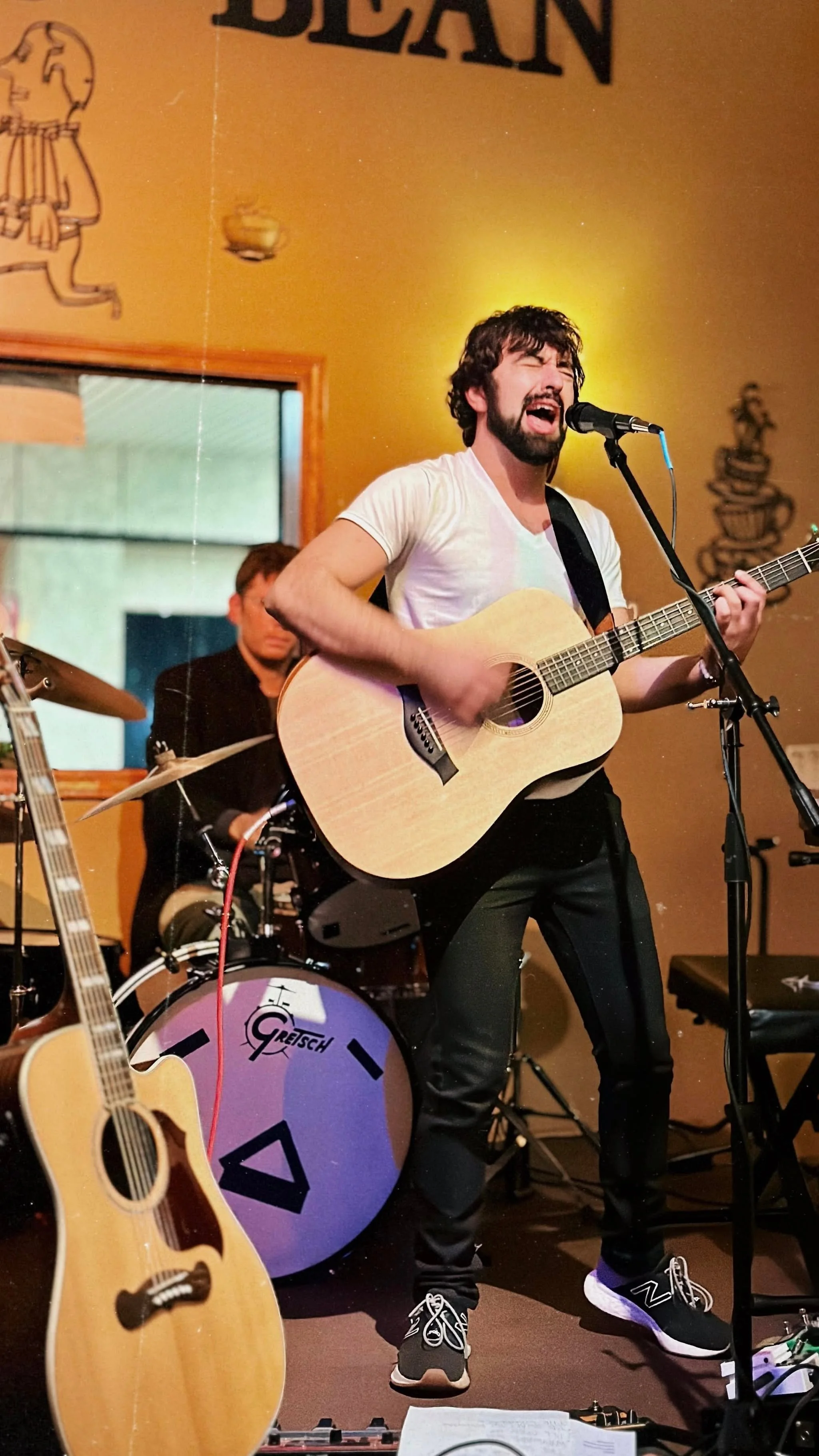 A musician with dark curly hair and beard singing passionately into a microphone while playing an acoustic guitar during a live performance. Behind him, a drummer with glasses sits at a drum set, and part of another guitar is visible in the foreground. The setting appears to be a cozy venue with warm lighting and wall art.