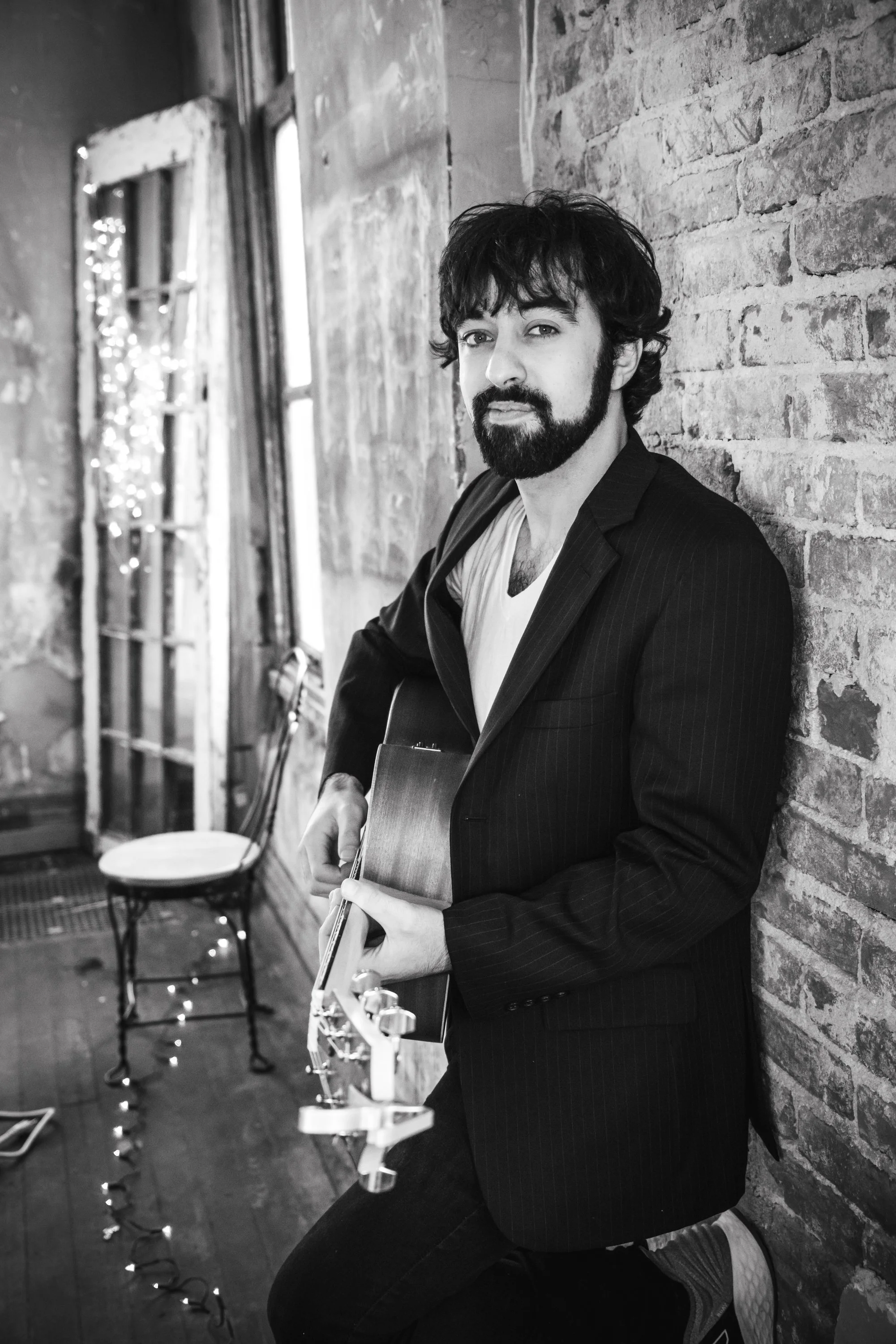 Joey Crofoot, wearing a black blazer and white shirt, is sitting against an exposed brick wall in a room with windows, holding a guitar and looking at the camera. The room has string lights, a chair, and some musical equipment.