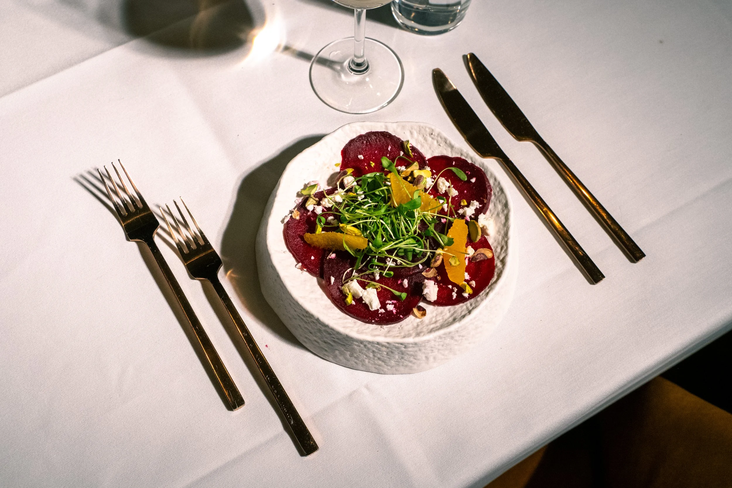 Ein Teller mit einer bunten Salatportion, garniert mit verschiedenen Sprossen, gerösteten Nüssen und streichfahigem Käse, auf einem weißen Tisch, begleitet von einem Weinglas, Messer und Gabel.