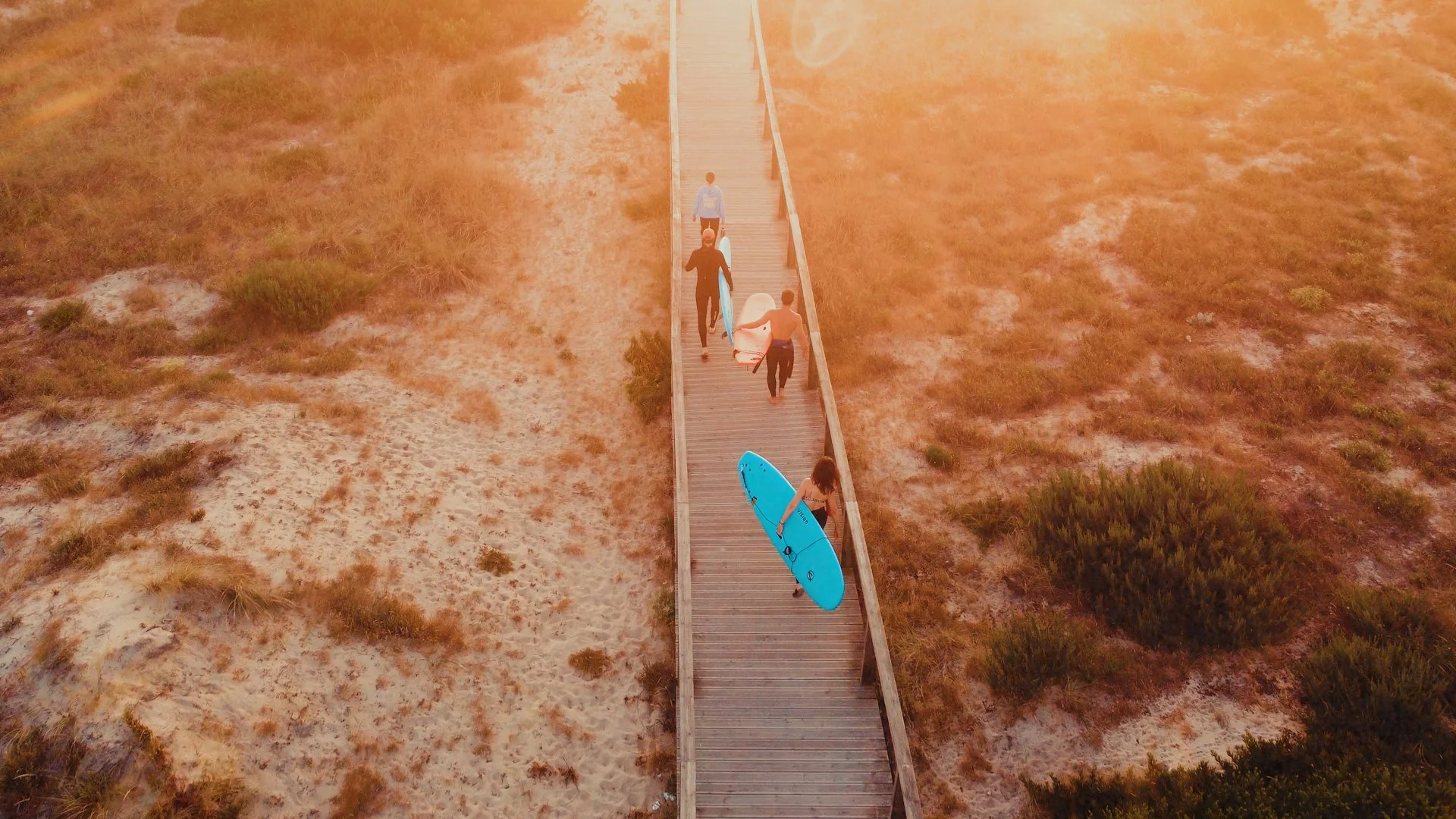 Vier Menschen gehen über eine längliche Holzbrücke am Strand, einer trägt ein Surfbrett, ein weiterer ist mit einem Surfboard unterwegs.