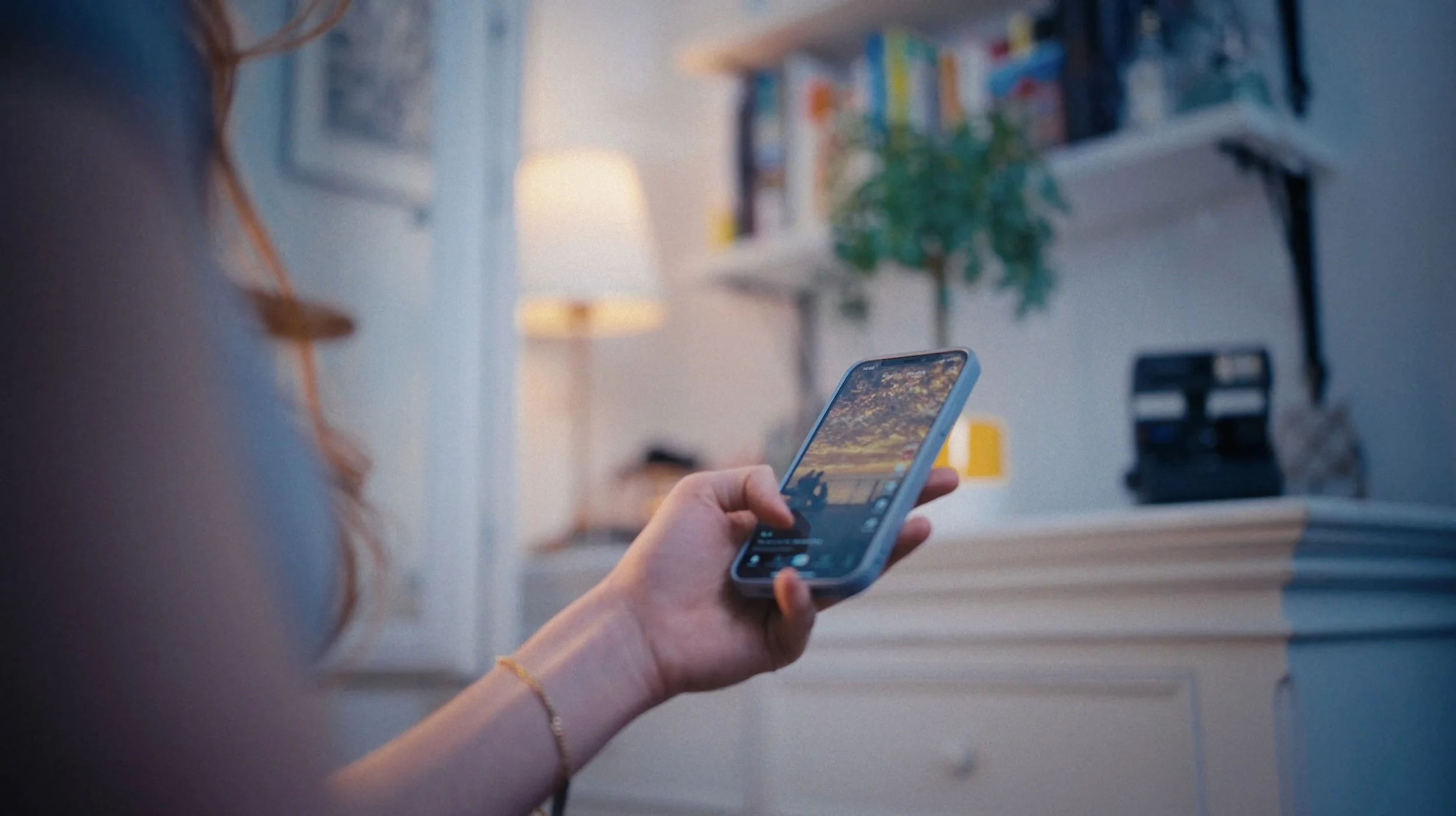 Person hält ein Smartphone in der Hand, während sie eine Fotobearbeitungs- oder Videobearbeitungs-App auf dem Bildschirm verwendet. Im Hintergrund befindet sich eine Bücherwand, ein Tisch mit einer Lampe und einigen Gegenständen, und es herrscht eine gemütliche Wohnzimmeratmosphäre.