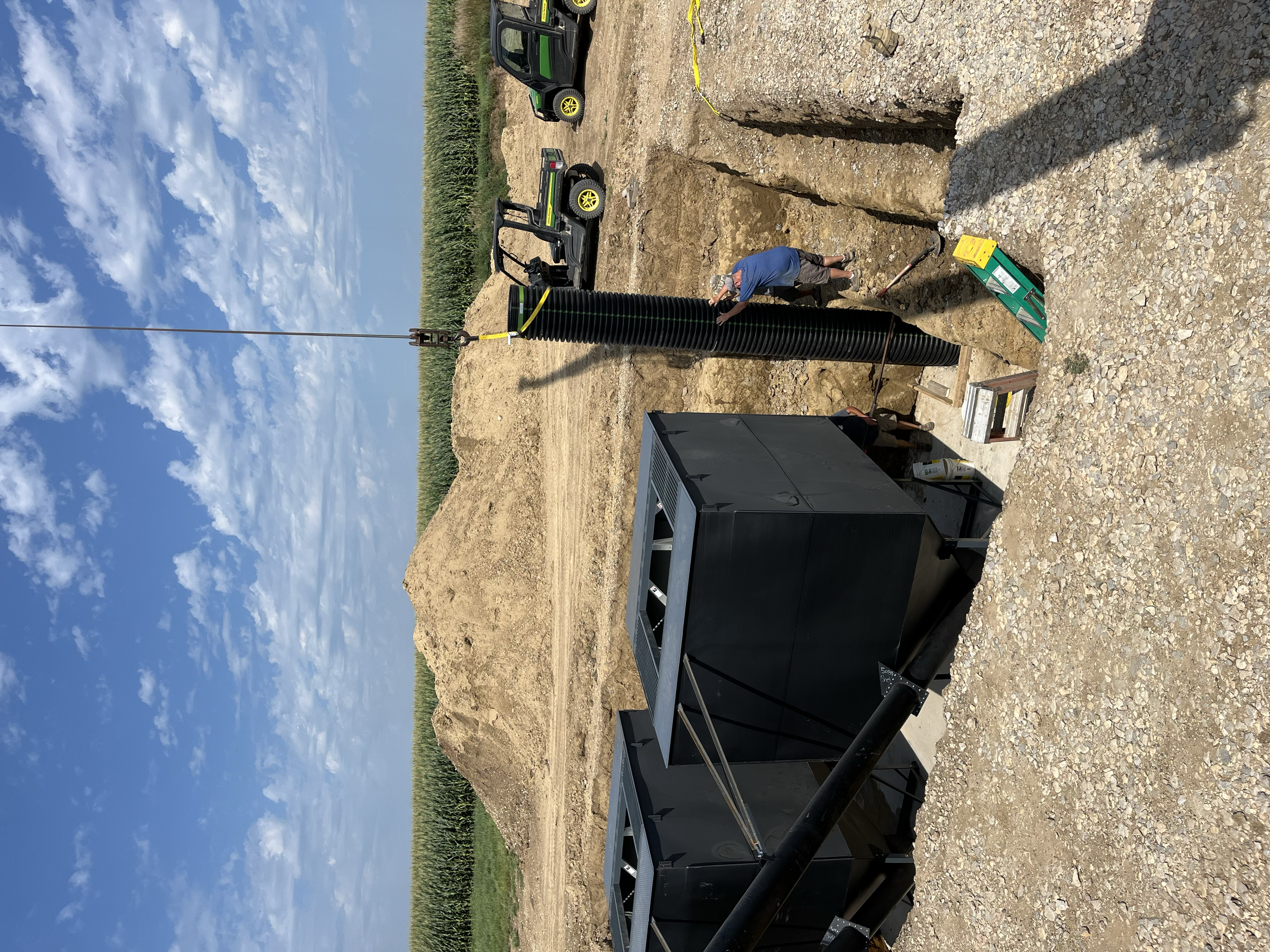 A worker crouching beside a large black pipe at an excavation site, with construction equipment and vehicles in the background, and a clear blue sky overhead.