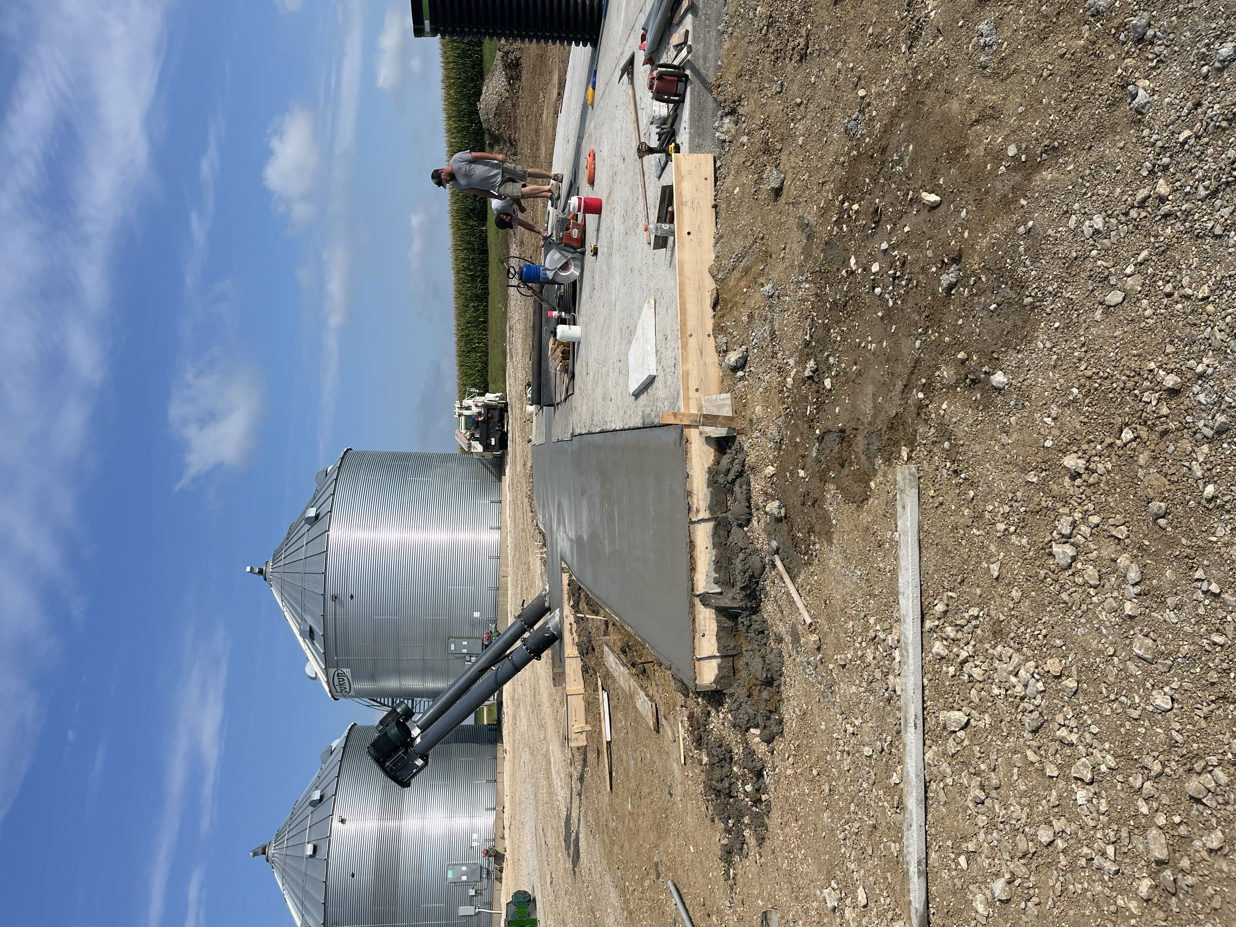 Construction workers pouring a concrete slab on a farm with large silos in the background.