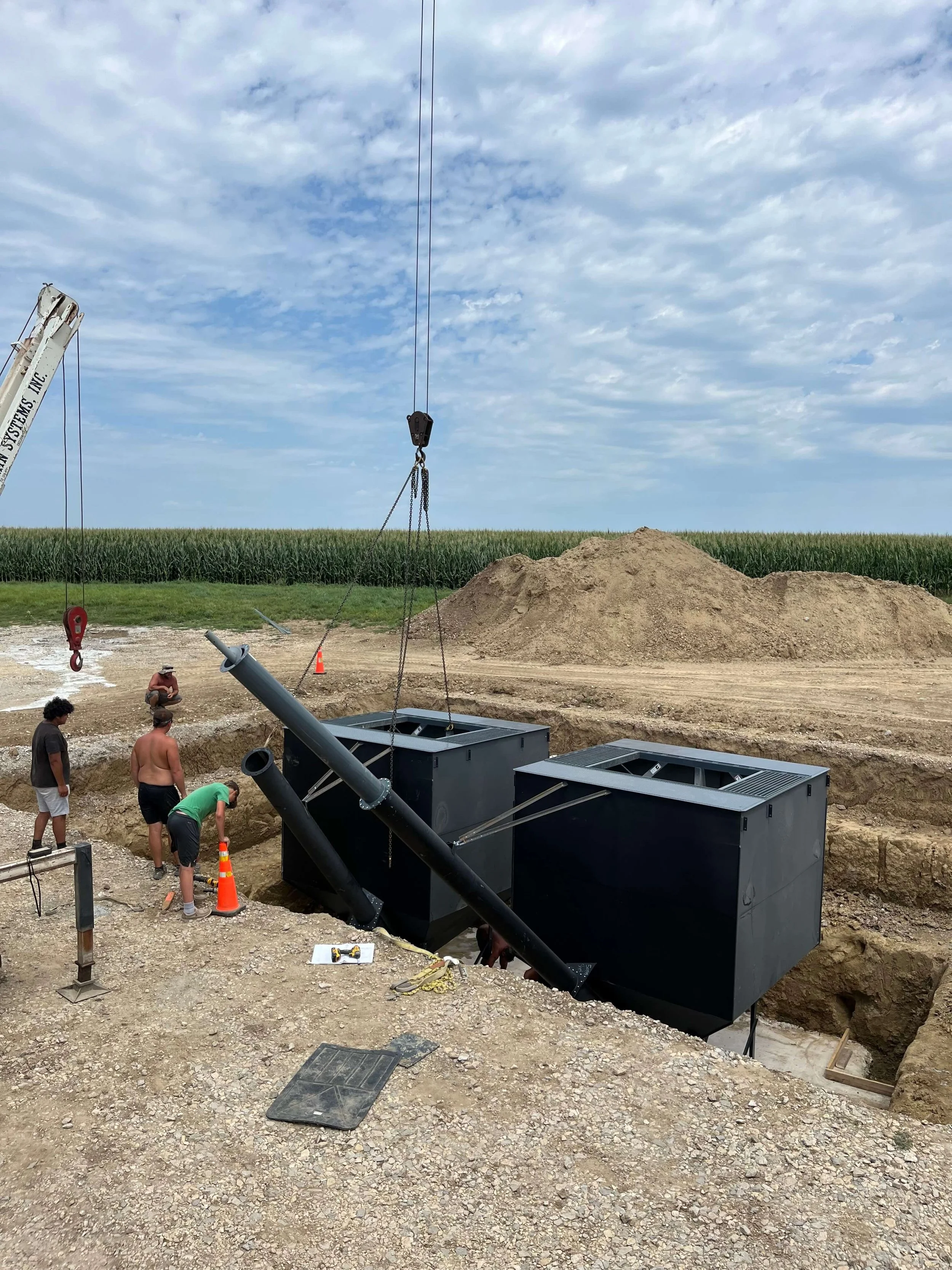 Construction workers installing a large underground utility box at a construction site with a cornfield in the background and a partly cloudy sky.