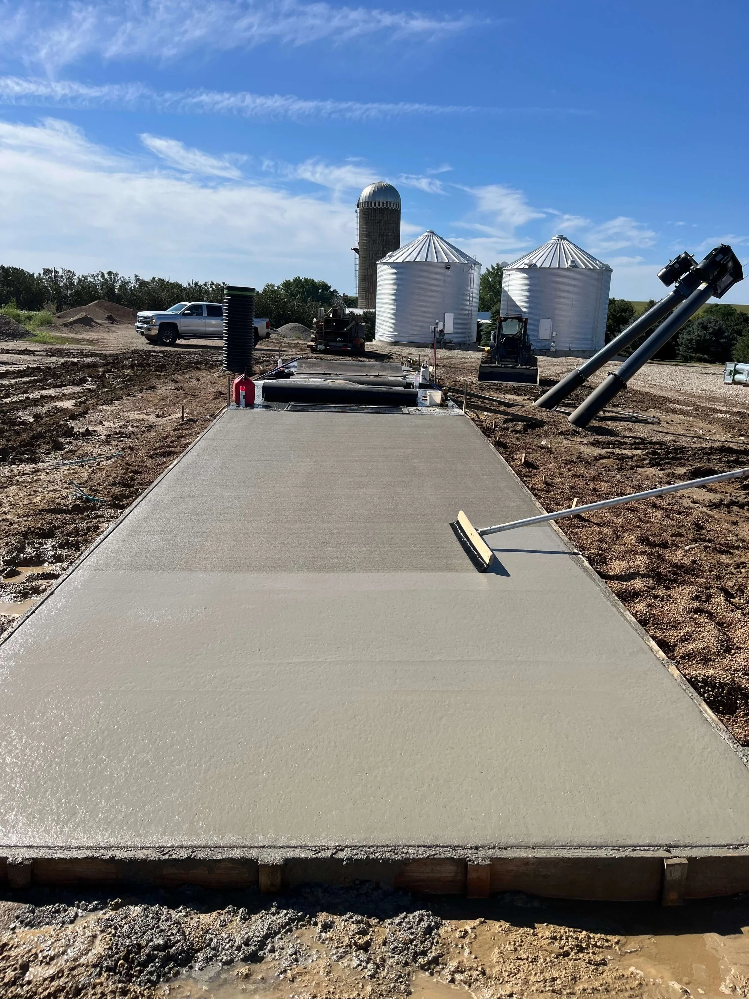 Freshly poured concrete on a building site with construction equipment, silos in the background, and a bright blue sky.