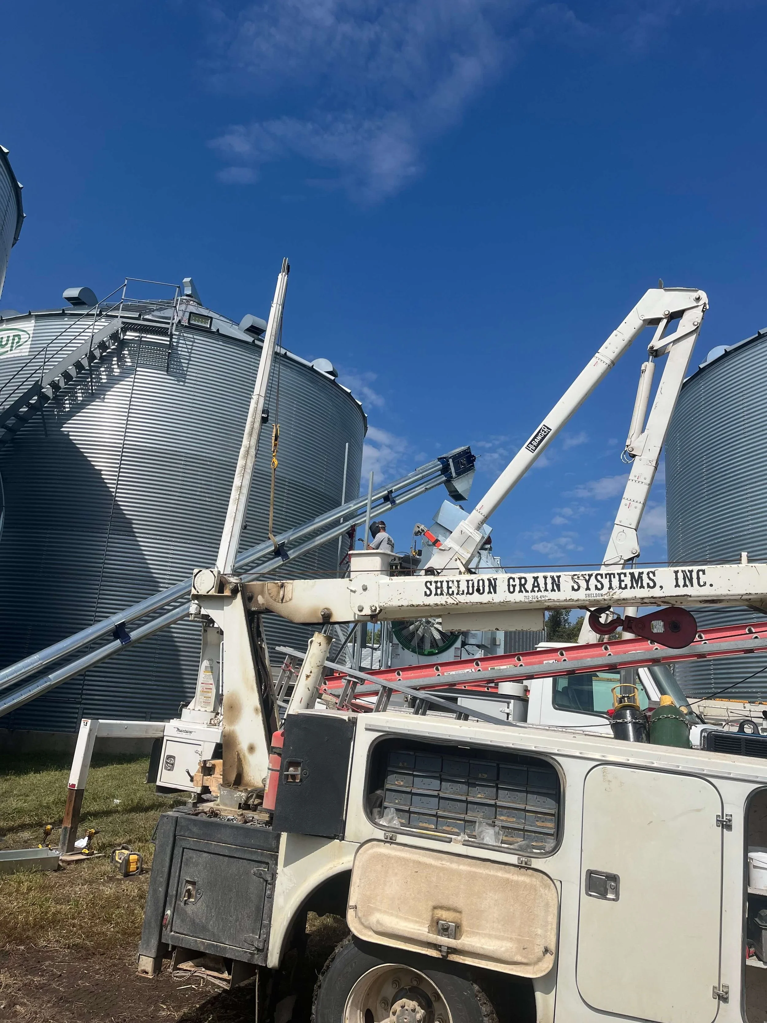 A utility truck with equipment and tools parked outdoors beneath a blue sky, with large grain storage silos in the background. Two workers are present, one on a crane platform working near the silos.