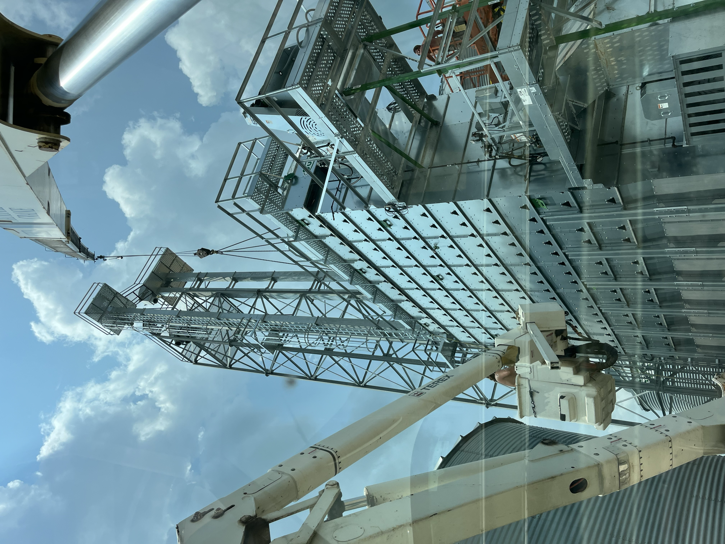 A close-up of construction equipment and scaffolding against a partly cloudy sky, with pipes, metal structures, and a crane with cables visible.