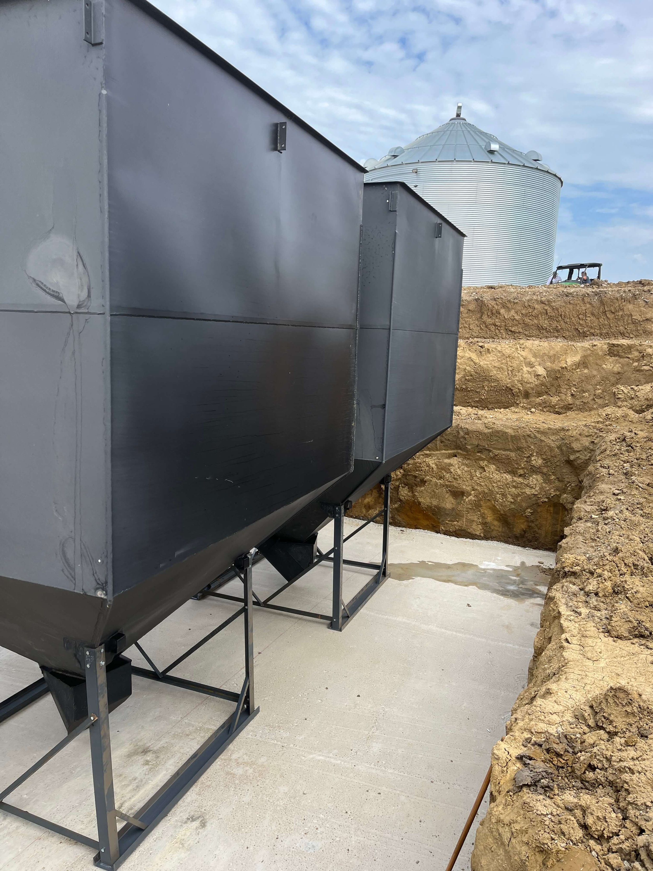 Large metal grain silo and utility shed on a construction site with exposed dirt and rocks, and a bulldozer in the background under a partly cloudy sky.