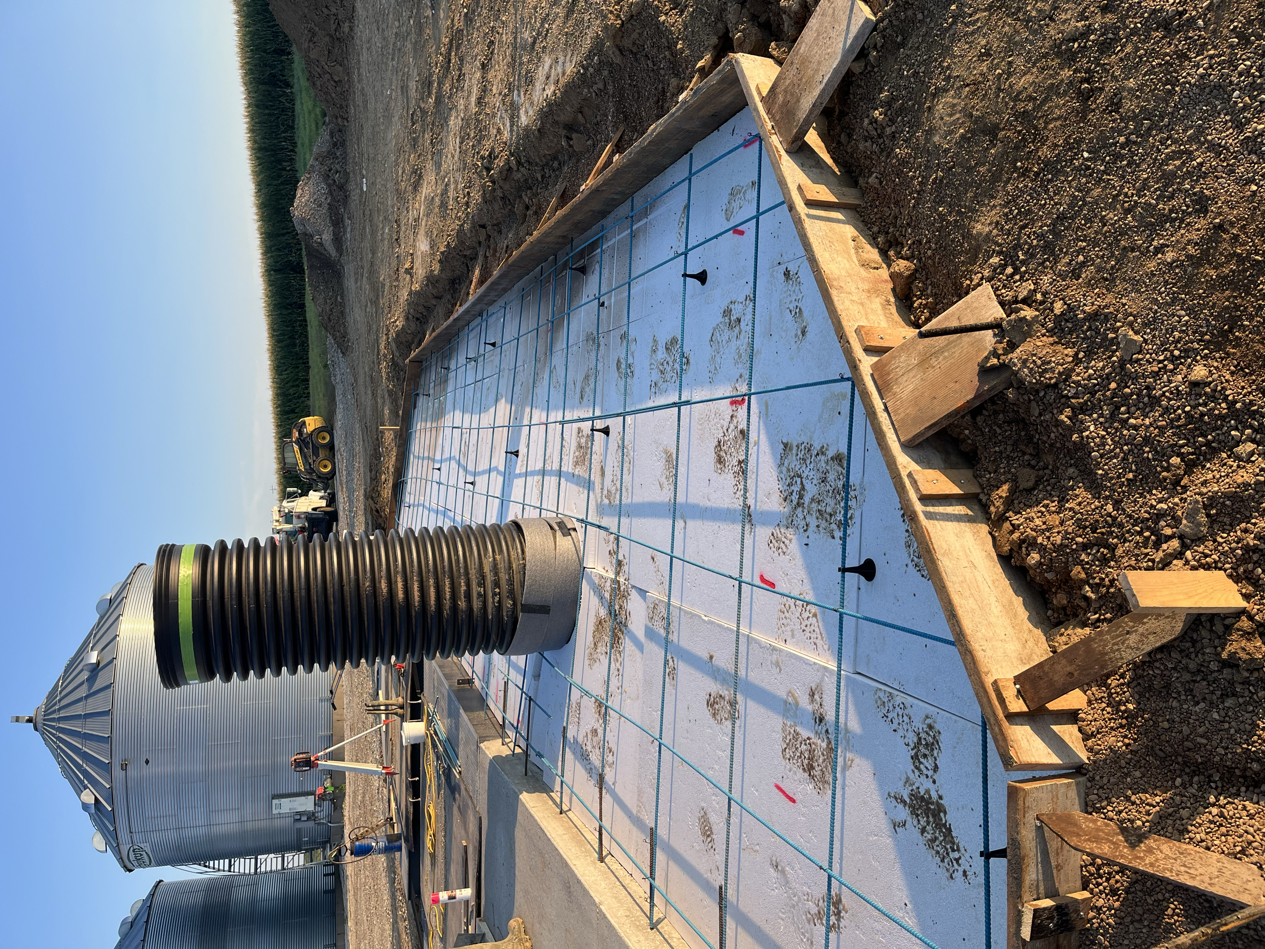 Construction site with concrete foundation, rebar, and nearby large metal grain silos under a clear blue sky.