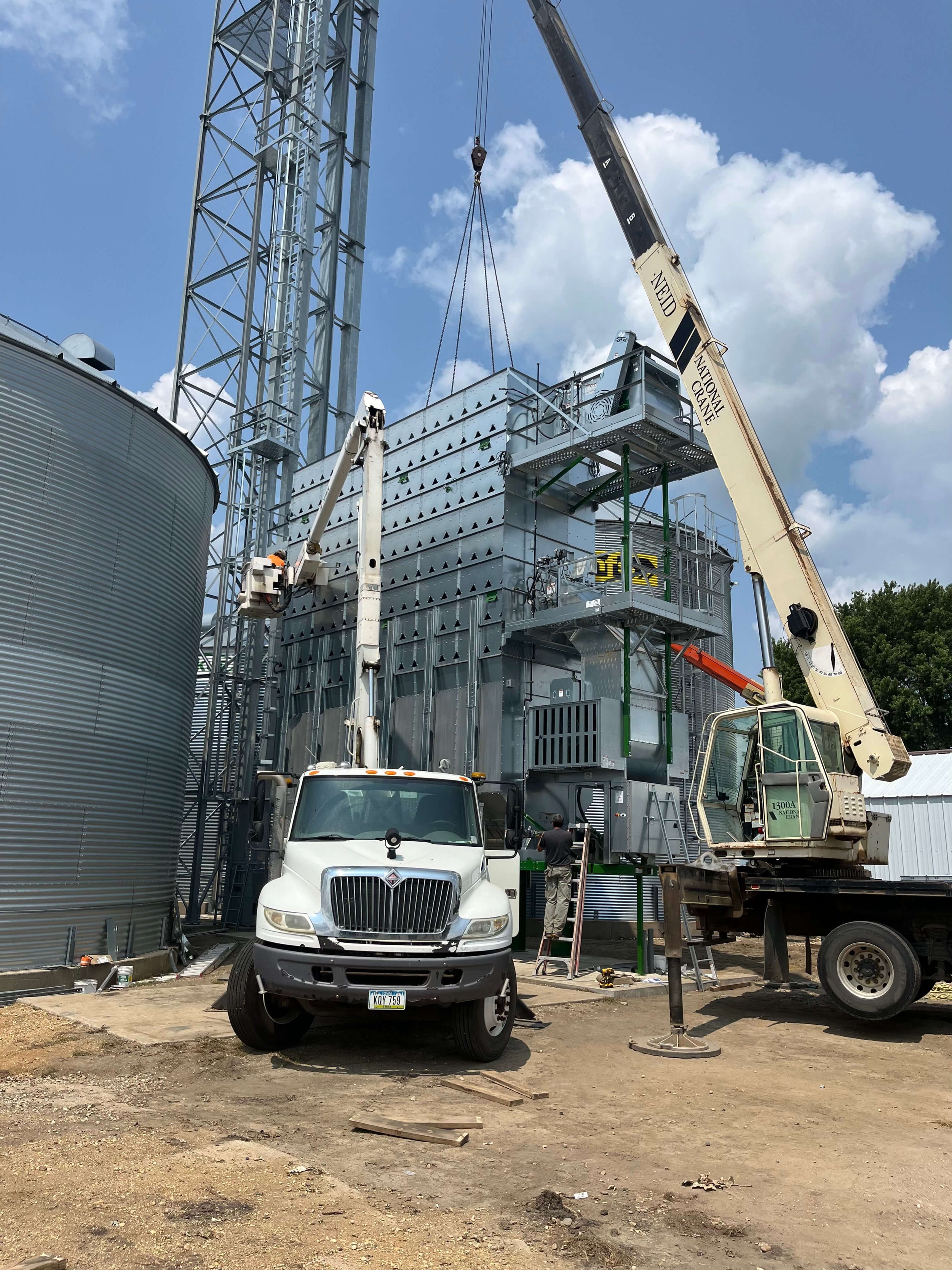 Construction site with a large metal structure, a crane lifting a section, a white truck and a worker on a ladder, and a dirt ground with scattered wood planks and debris.