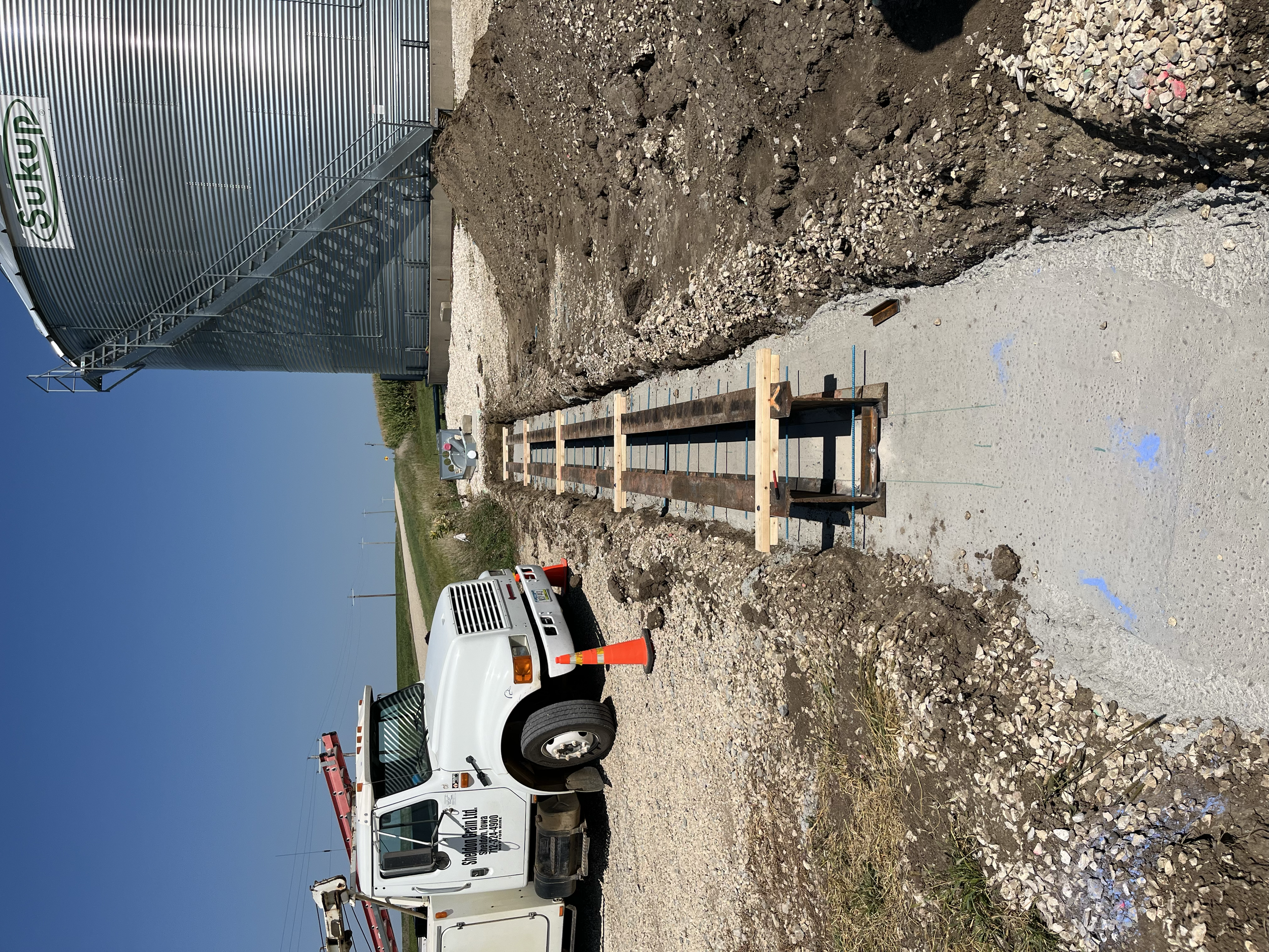 Construction site with a white utility truck and a large pile of dirt and gravel. The truck has a red ladder on top and appears to be used for construction or landscaping work. In the background, there is a metal building with a staircase leading to 