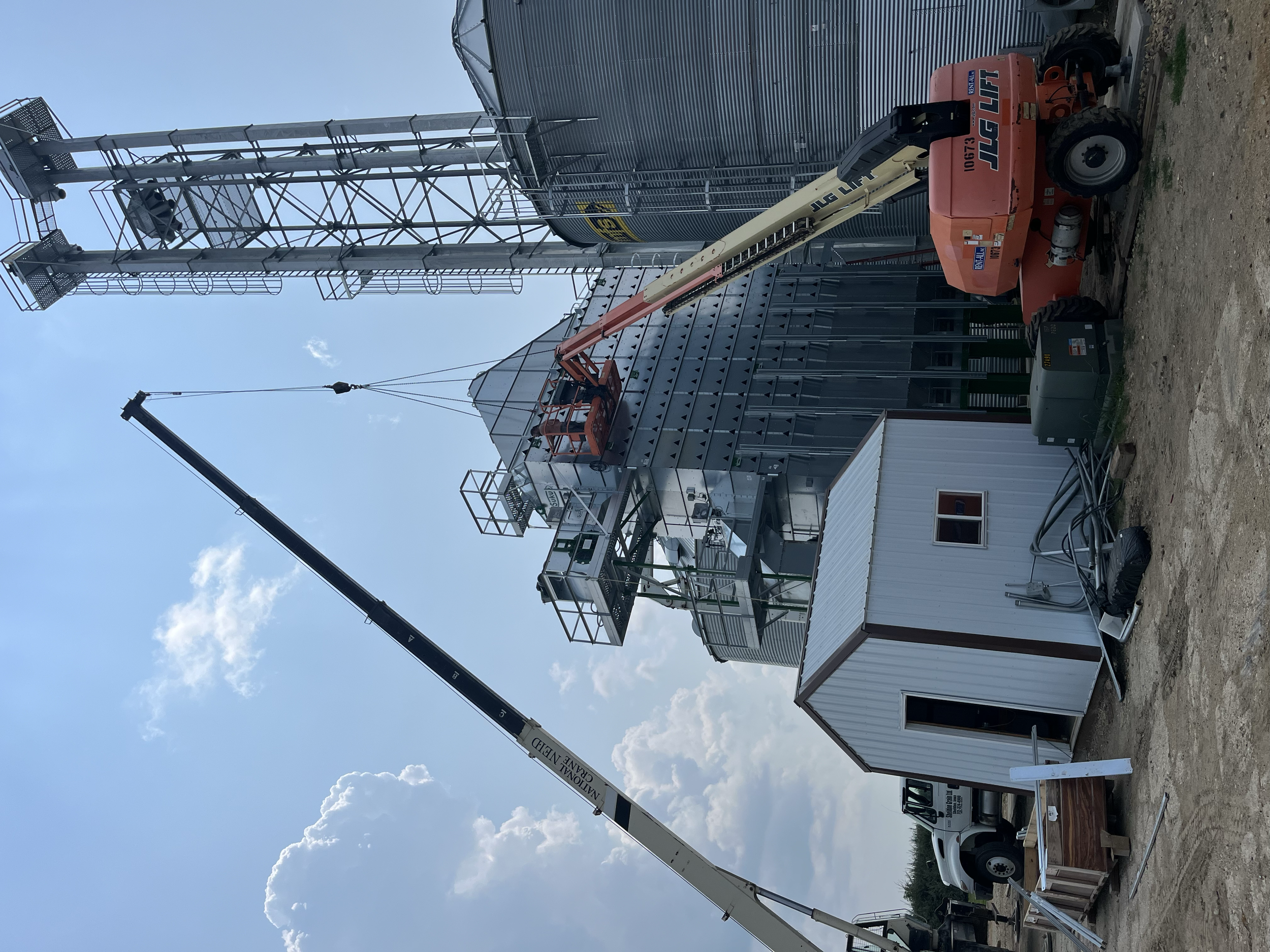 Construction site featuring a tall metal tower with a platform and ladders, a large metal building, construction vehicles, including a cherry picker, and various construction materials, under a partly cloudy sky.