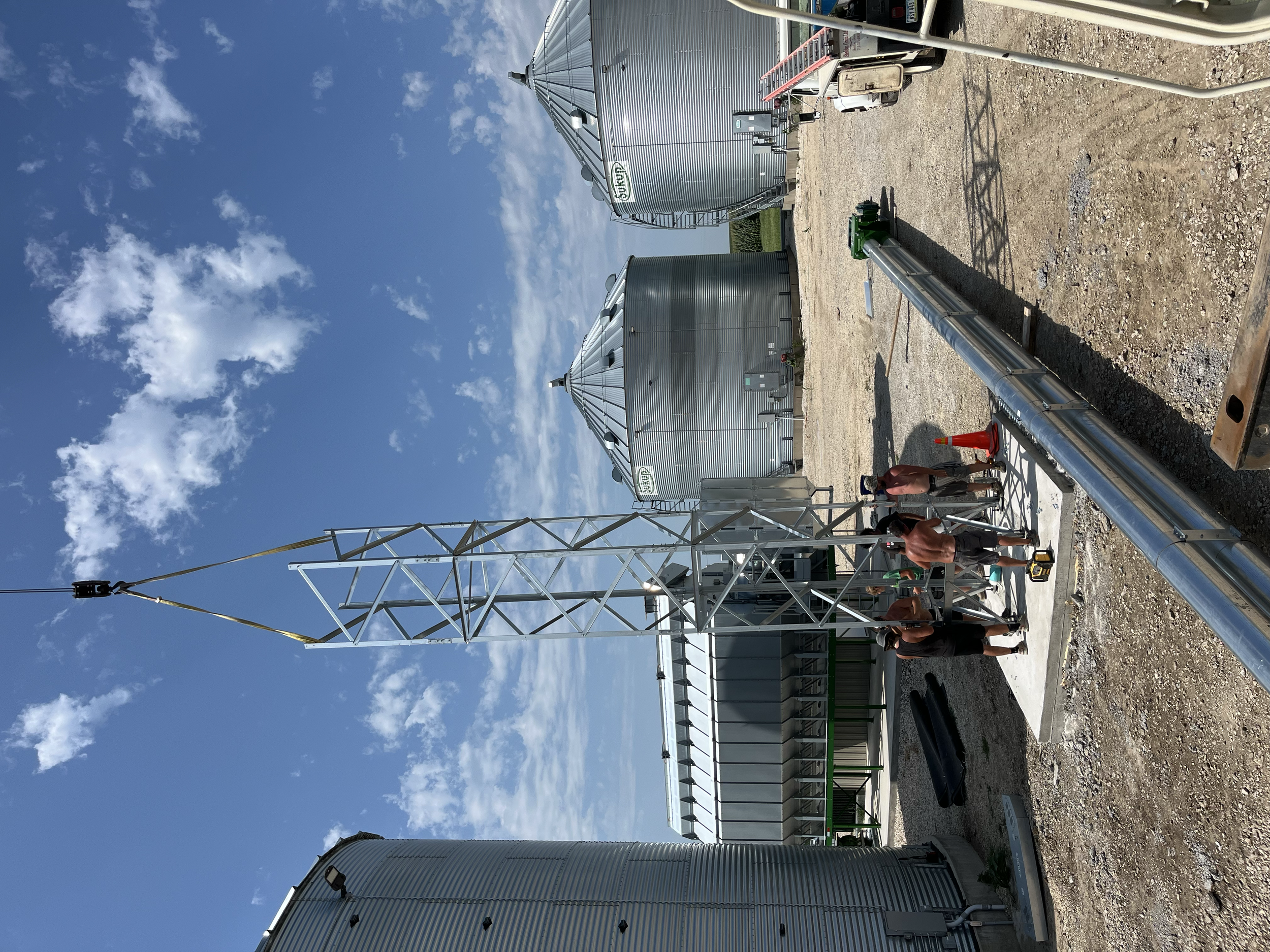 Three large metal grain storage silos in an outdoor setting on a sunny day with a clear blue sky and some clouds. Several people are working near the silos, and construction or maintenance equipment is present.