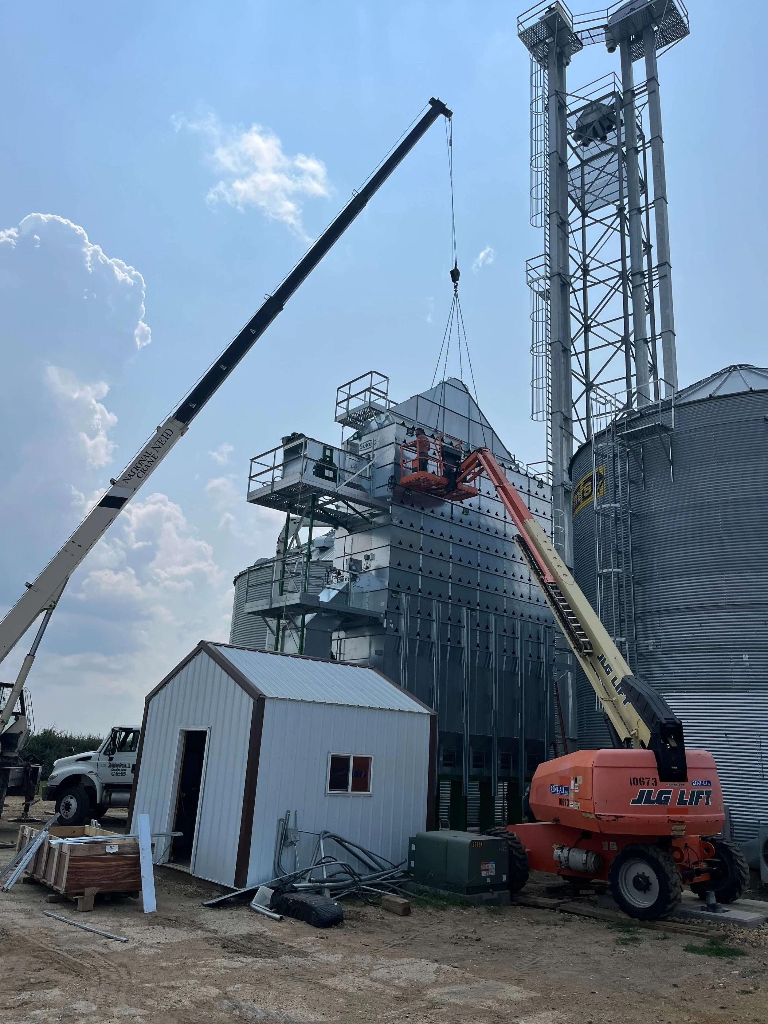 Construction site with a crane lifting a large metal tank to a grain silo. There is a small white building in the foreground, construction equipment, and a partly cloudy sky.