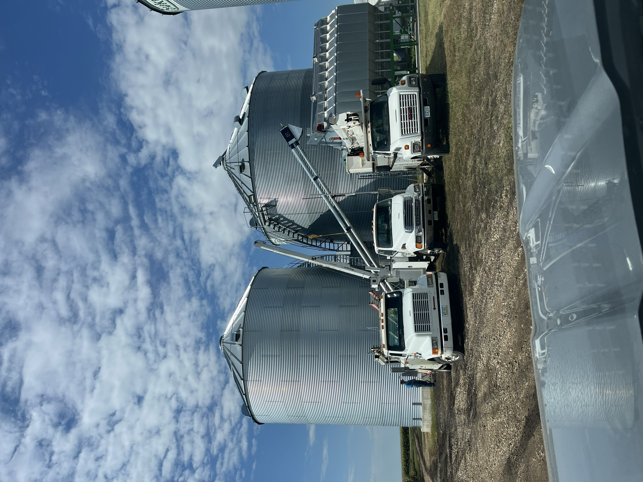 A white utility truck with an extended ladder and large cylindrical grain silos in the background, set against a partly cloudy sky.