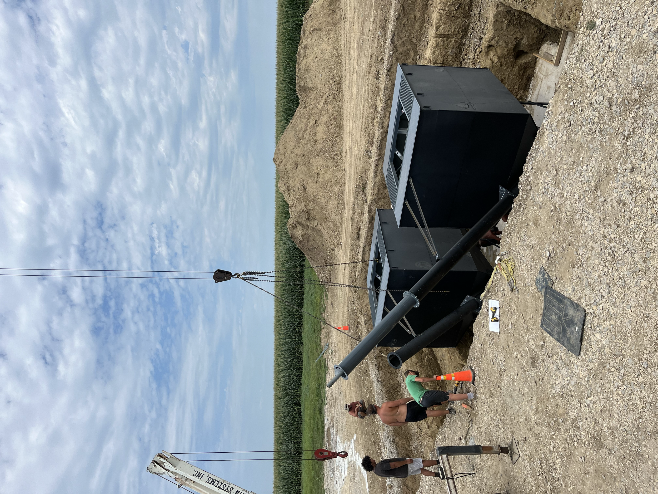 Construction site with large black pipe, workers, and safety equipment. Overcast sky, green field in background.