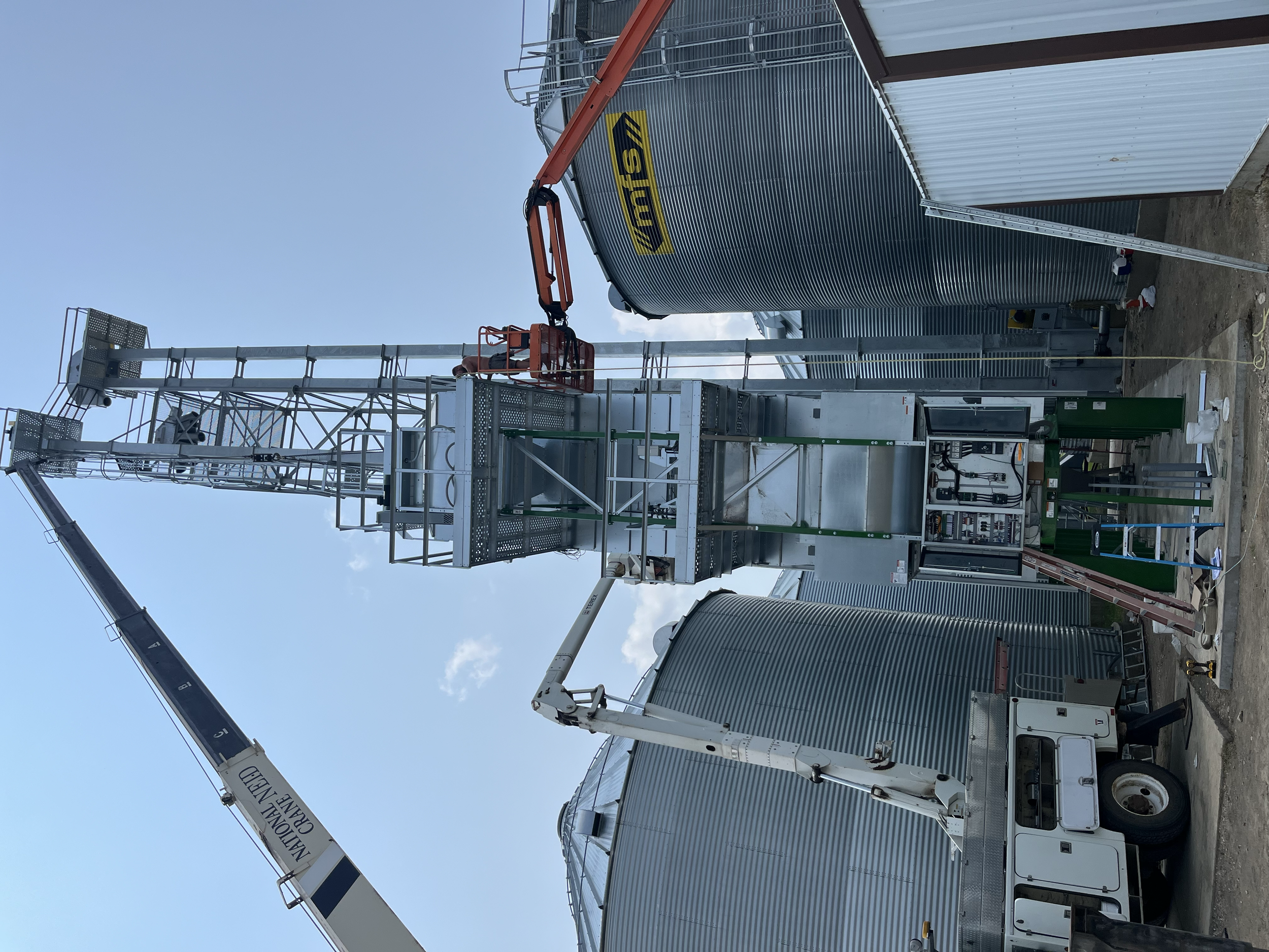 A large white truck with a crane lifting a silos in an industrial area, under a blue sky with some clouds.