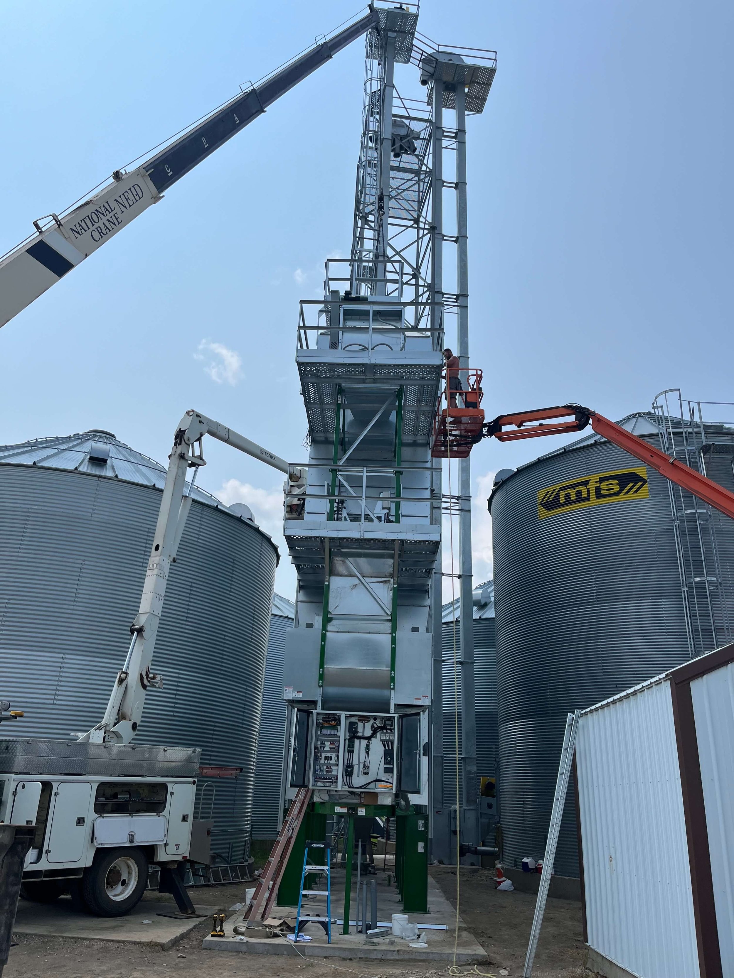 Construction site with a tall metal structure and a crane labeled 'National Need Crane' lifting equipment. Several large cylindrical metal grain silos are in the background, alongside various construction tools and materials on the ground.