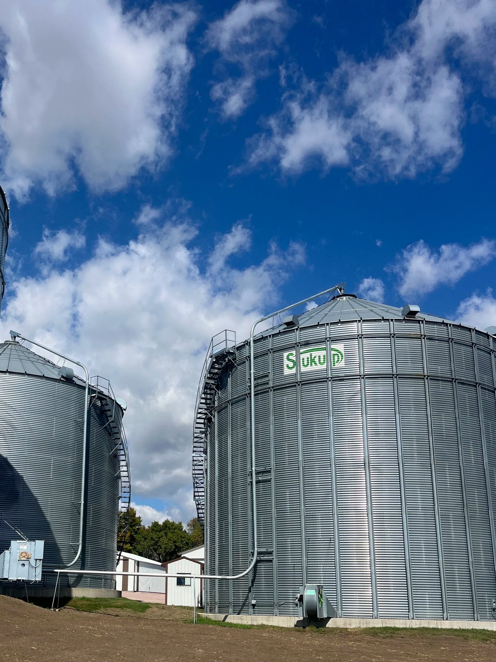 Large metal grain silos in an open area with a partly cloudy blue sky and some trees in the background.