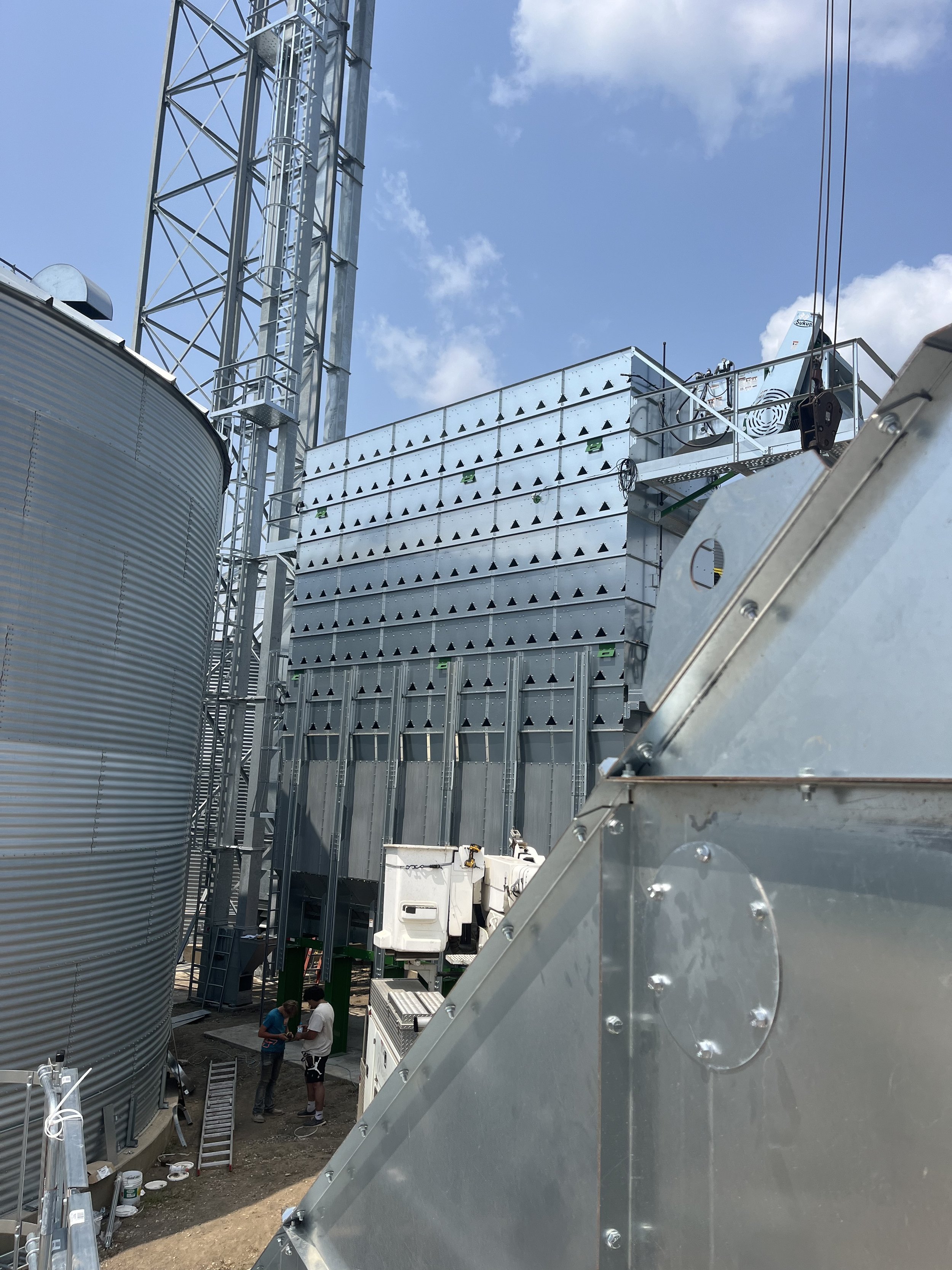 Two workers stand near commercial HVAC equipment, including large they are engaged in a discussion at an industrial site under a sunny sky.