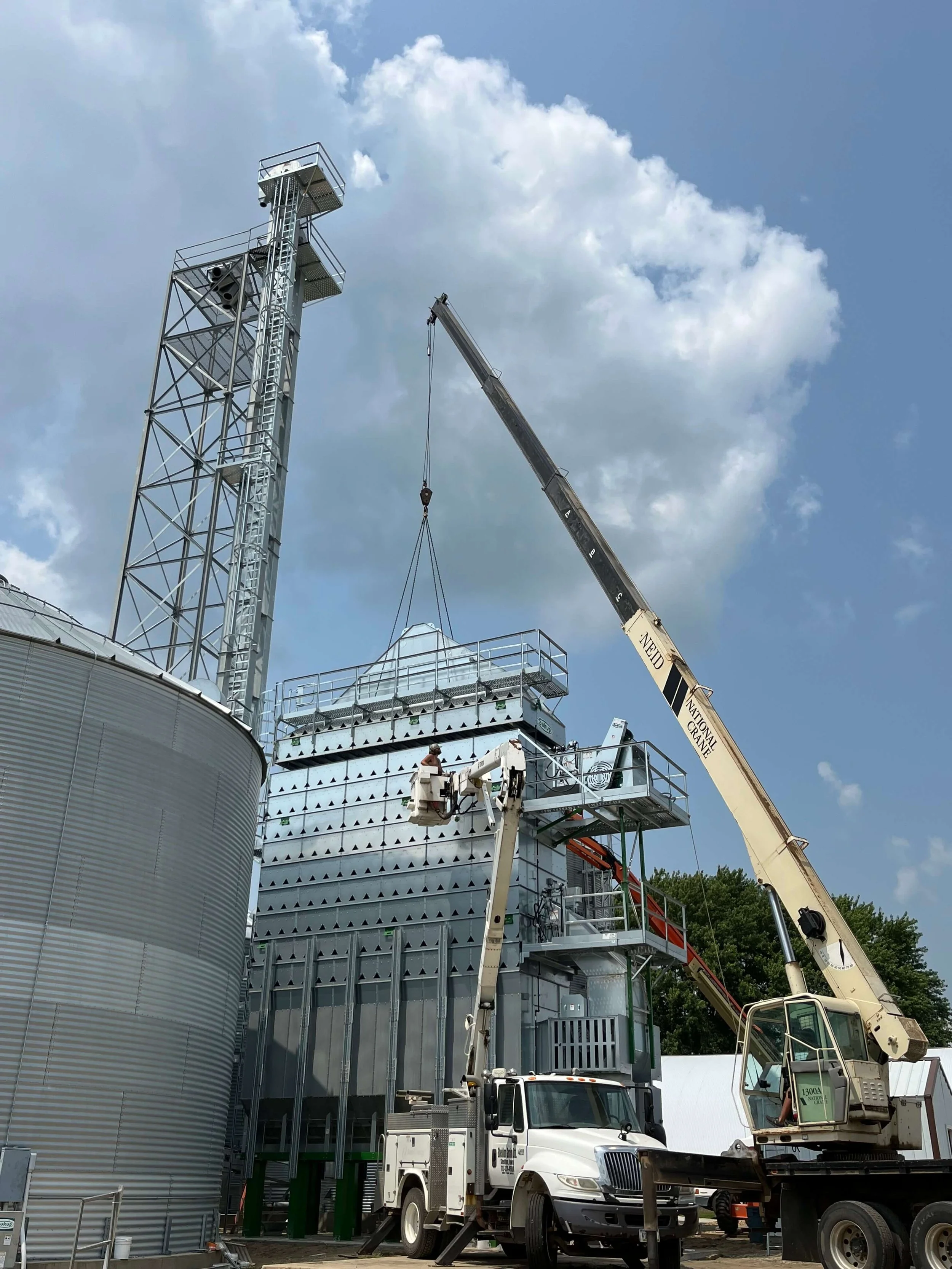 Construction workers operating crane on industrial site with metal structures, large storage tanks, and cloudy sky in the background.