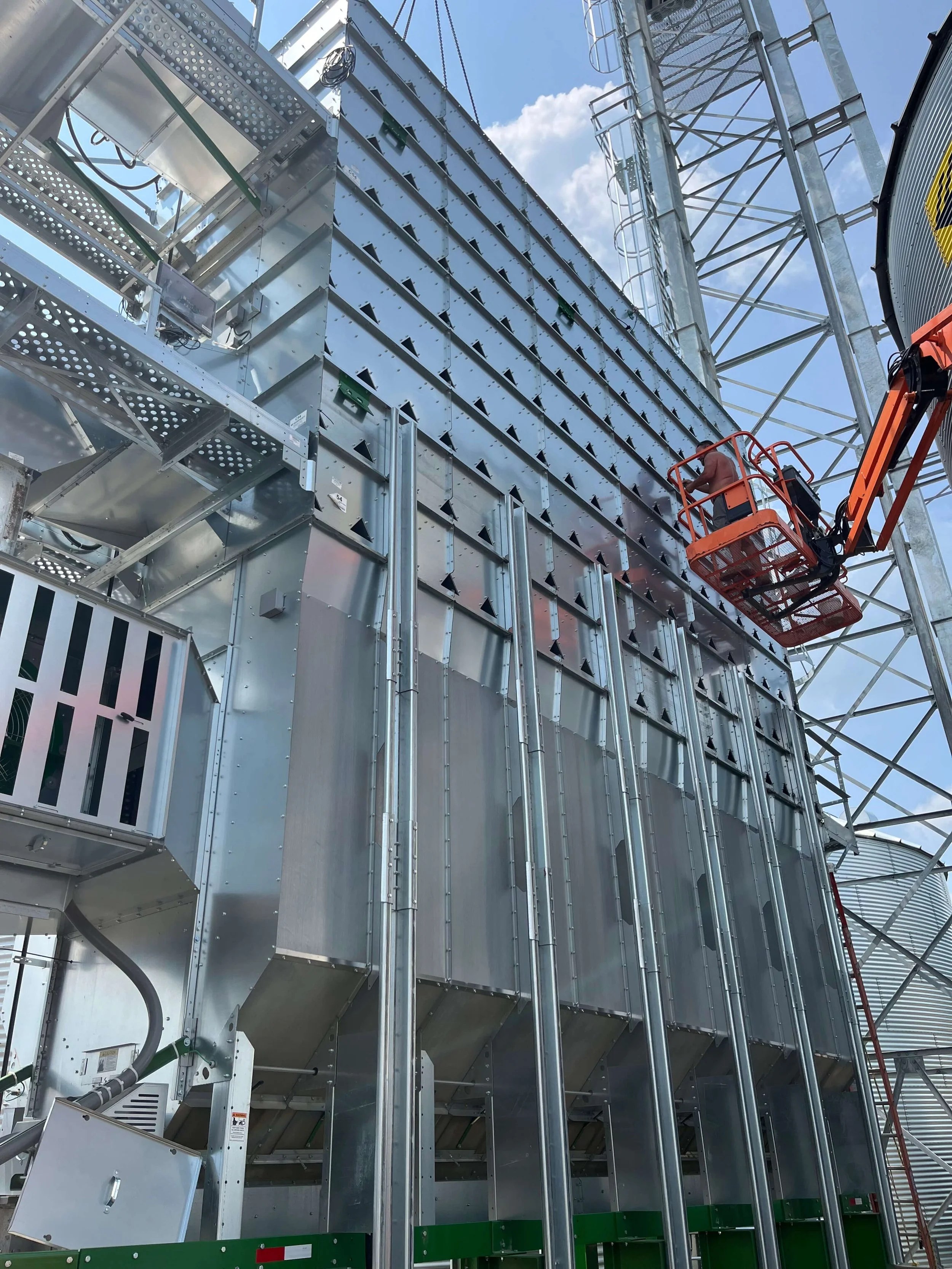 A construction worker is using a cherry picker lift to work on a large, industrial metal structure with vertical and horizontal beams. The background shows a blue sky with some clouds.