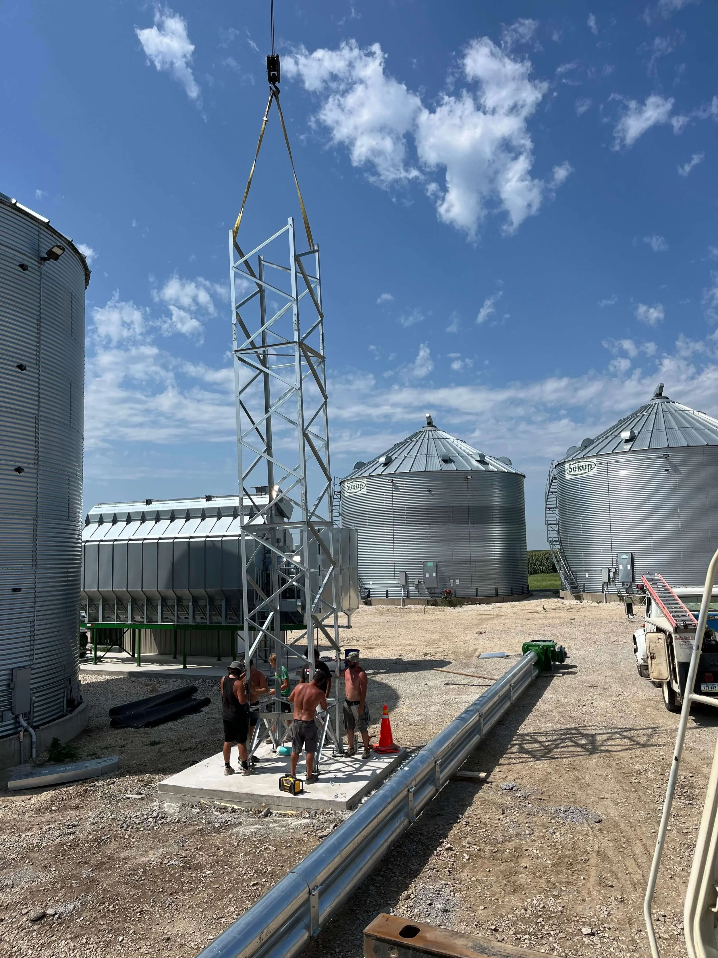 Workers installing a metal tower structure at an industrial site with large grain silos in the background, under a partly cloudy blue sky.