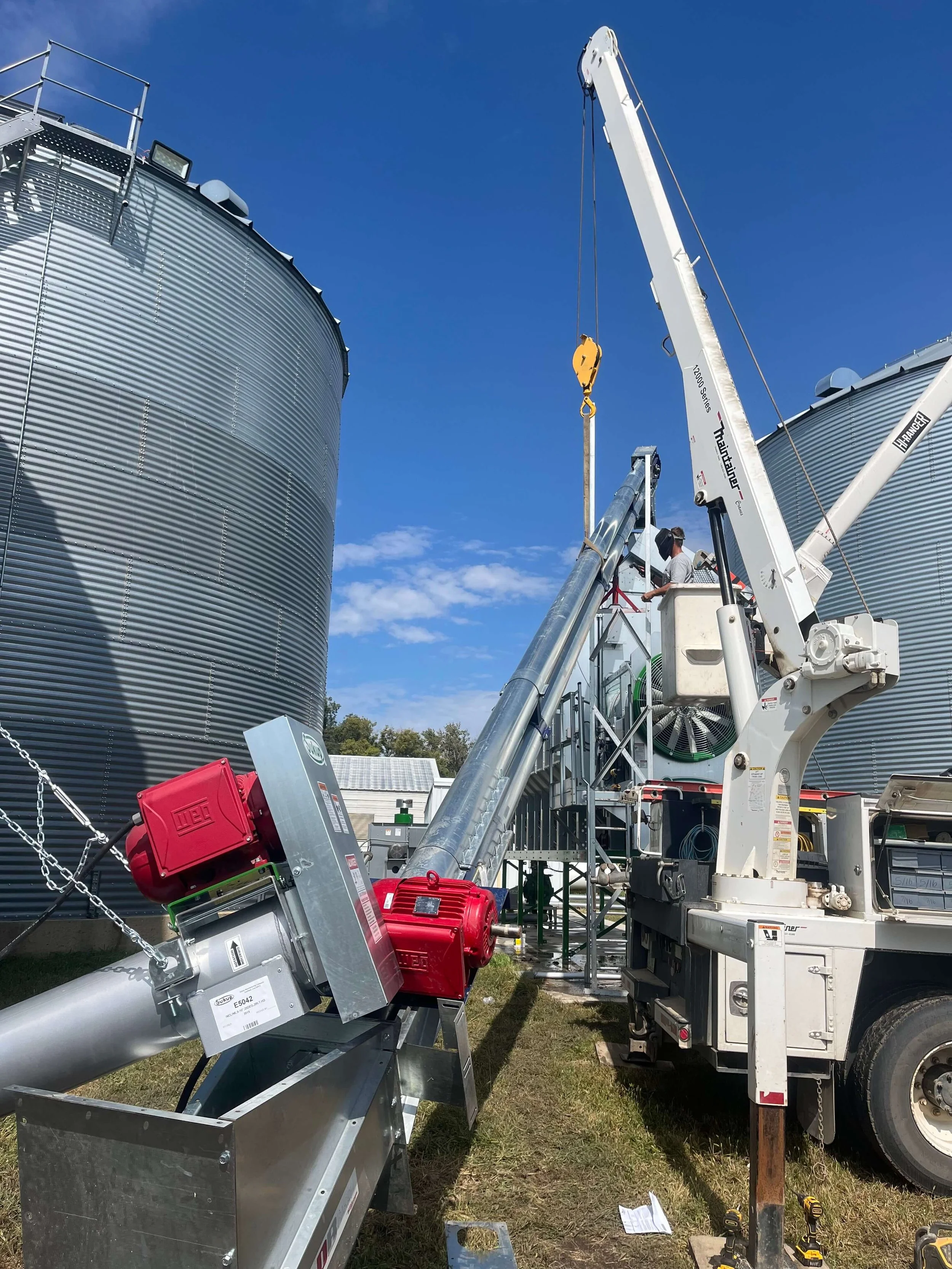Construction workers operating a crane on a farm with large metallic grain silos in the background, with a clear blue sky.