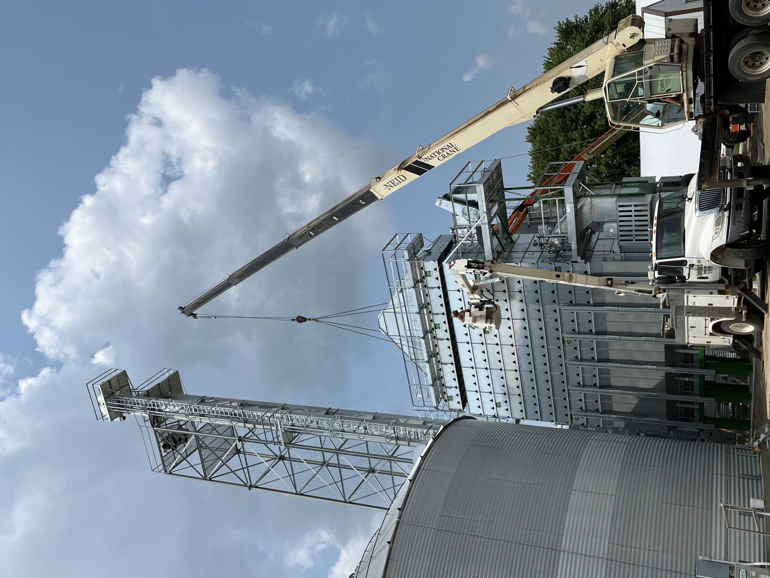 Construction crane lifting materials near large metal grain silos against a partly cloudy sky.