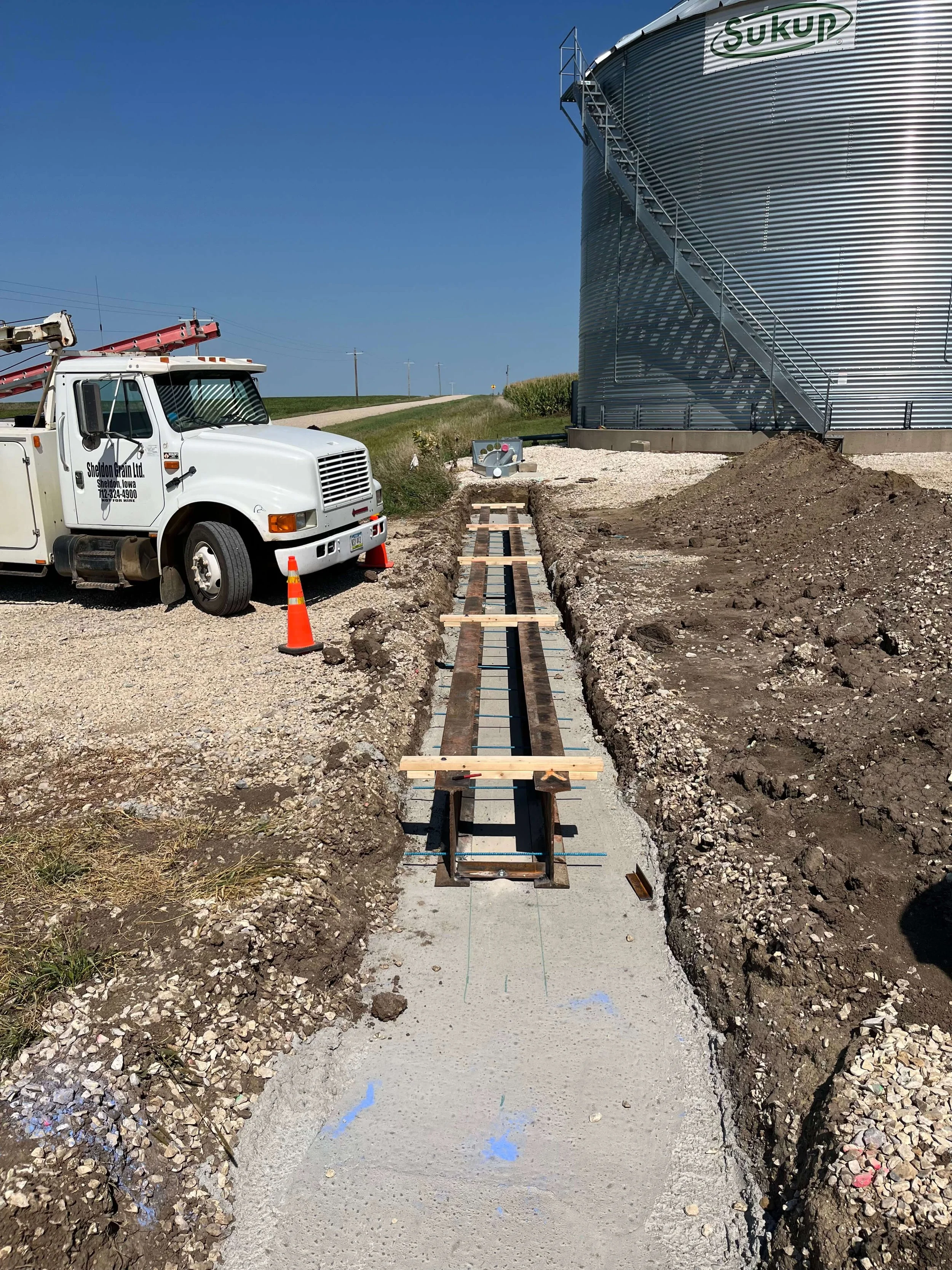 Construction site with a gravel pathway being built, a white utility truck with orange cones nearby, and a large metal water tank labeled "Sukup" in the background under a clear blue sky.